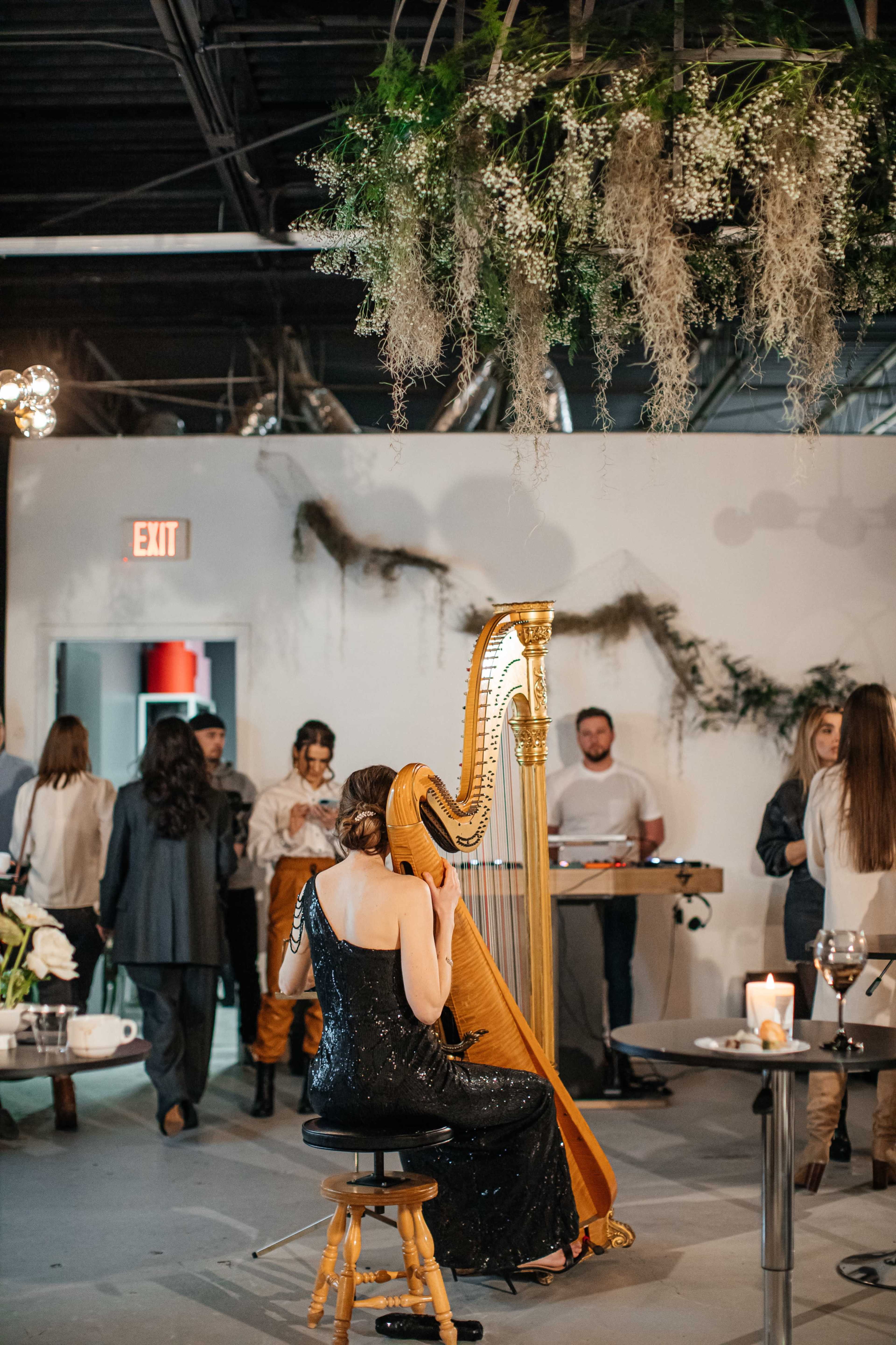 A woman in a black dress plays the harp in a decorated industrial space while guests mingle in the background.