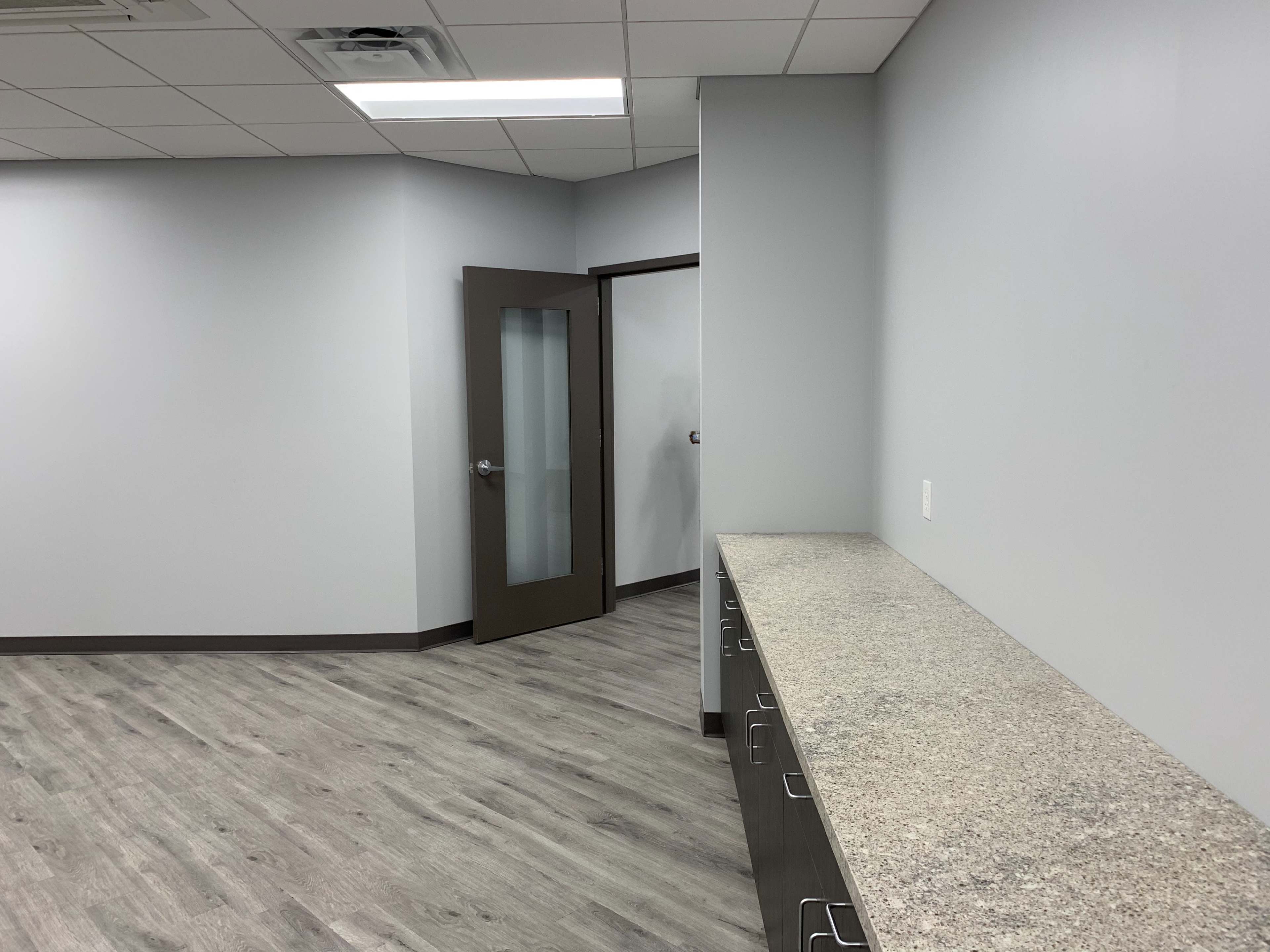 The image shows a modern, empty room with light gray walls, a long countertop along one wall, and a door leading to another room.