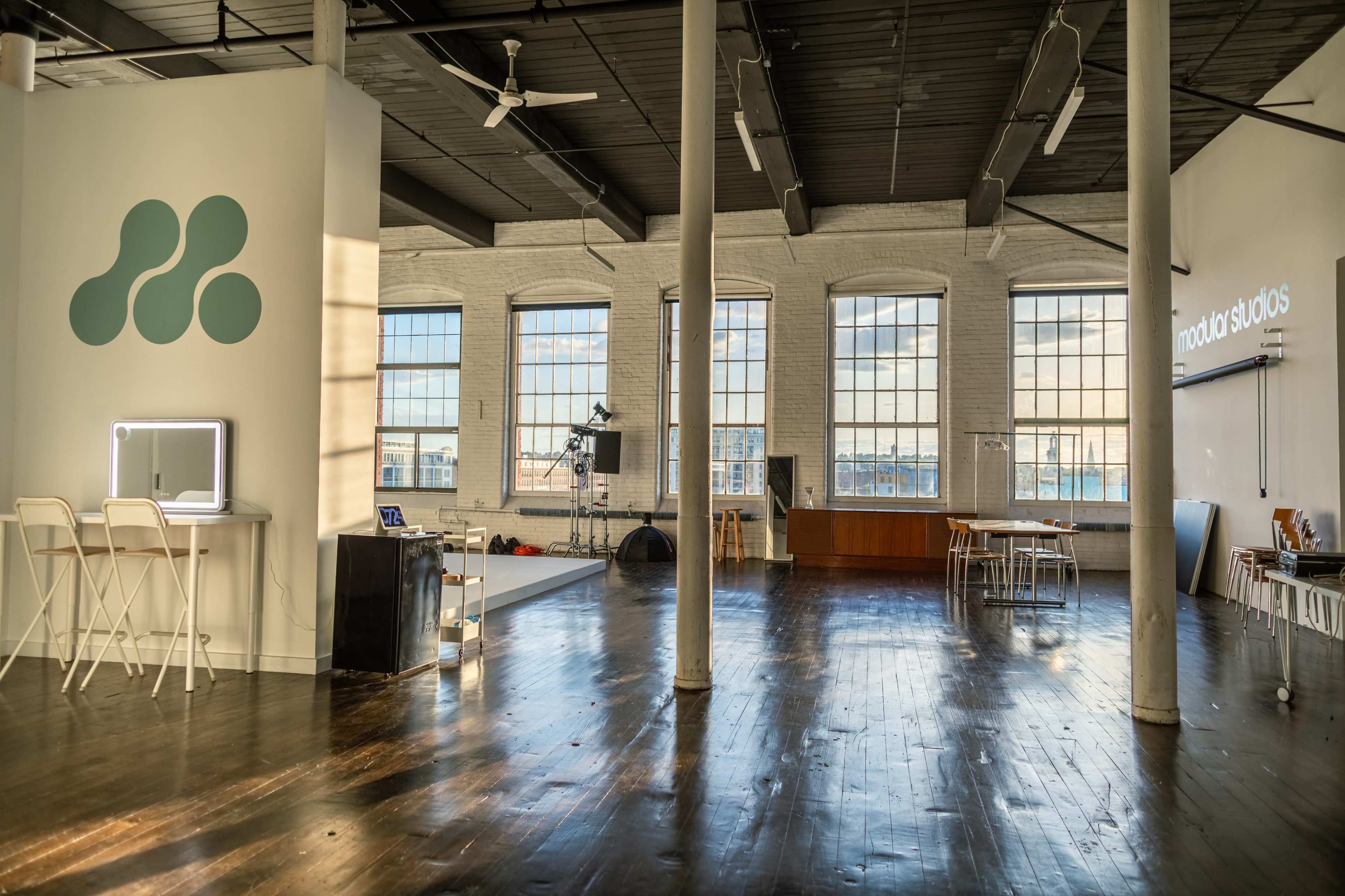 A spacious, well-lit studio with large windows, wooden flooring, and various furnishings including a table and chairs.