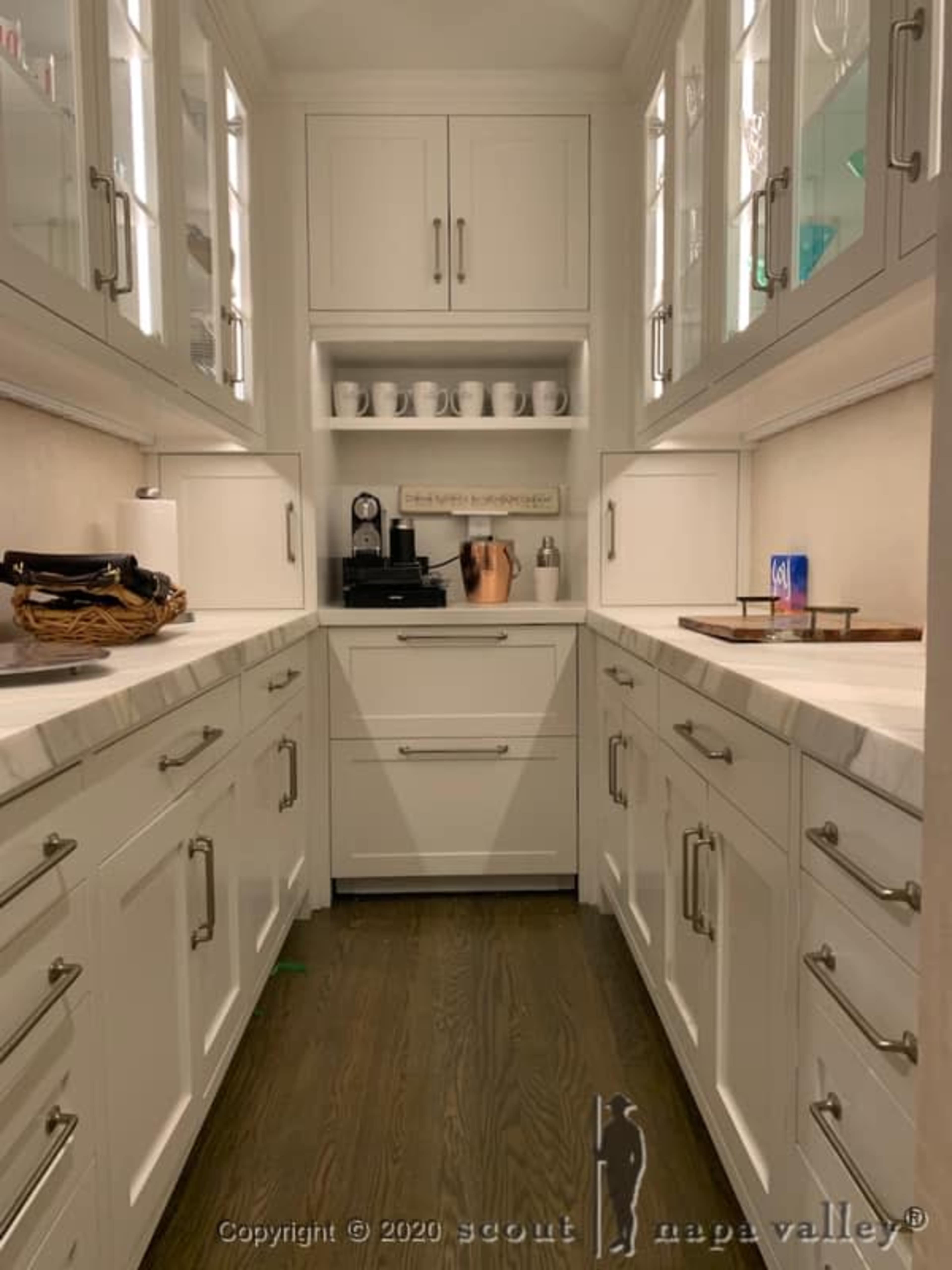 A narrow kitchen corridor with white cabinetry, marble countertops, and a coffee station arranged against the back wall.