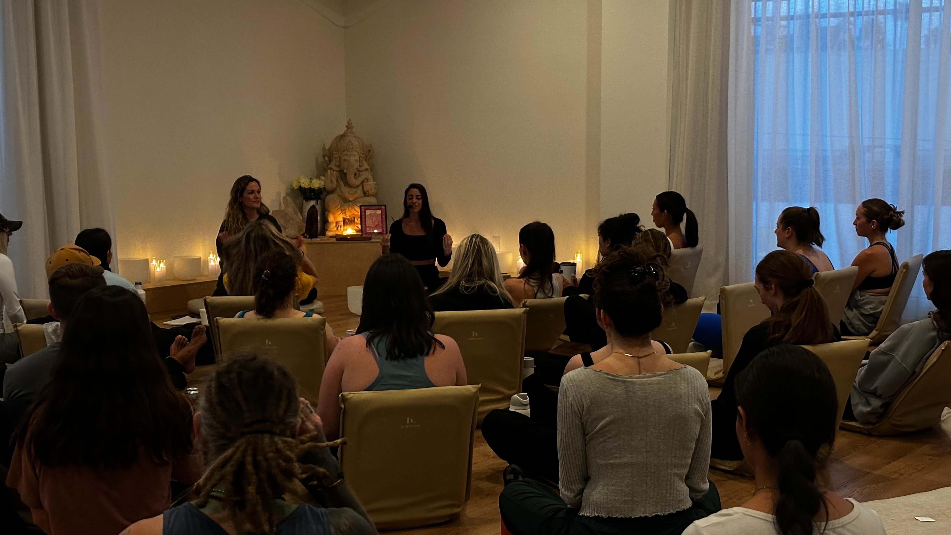 A group of individuals sits in a well-lit room, attentively facing two women who are speaking at the front near an altar.