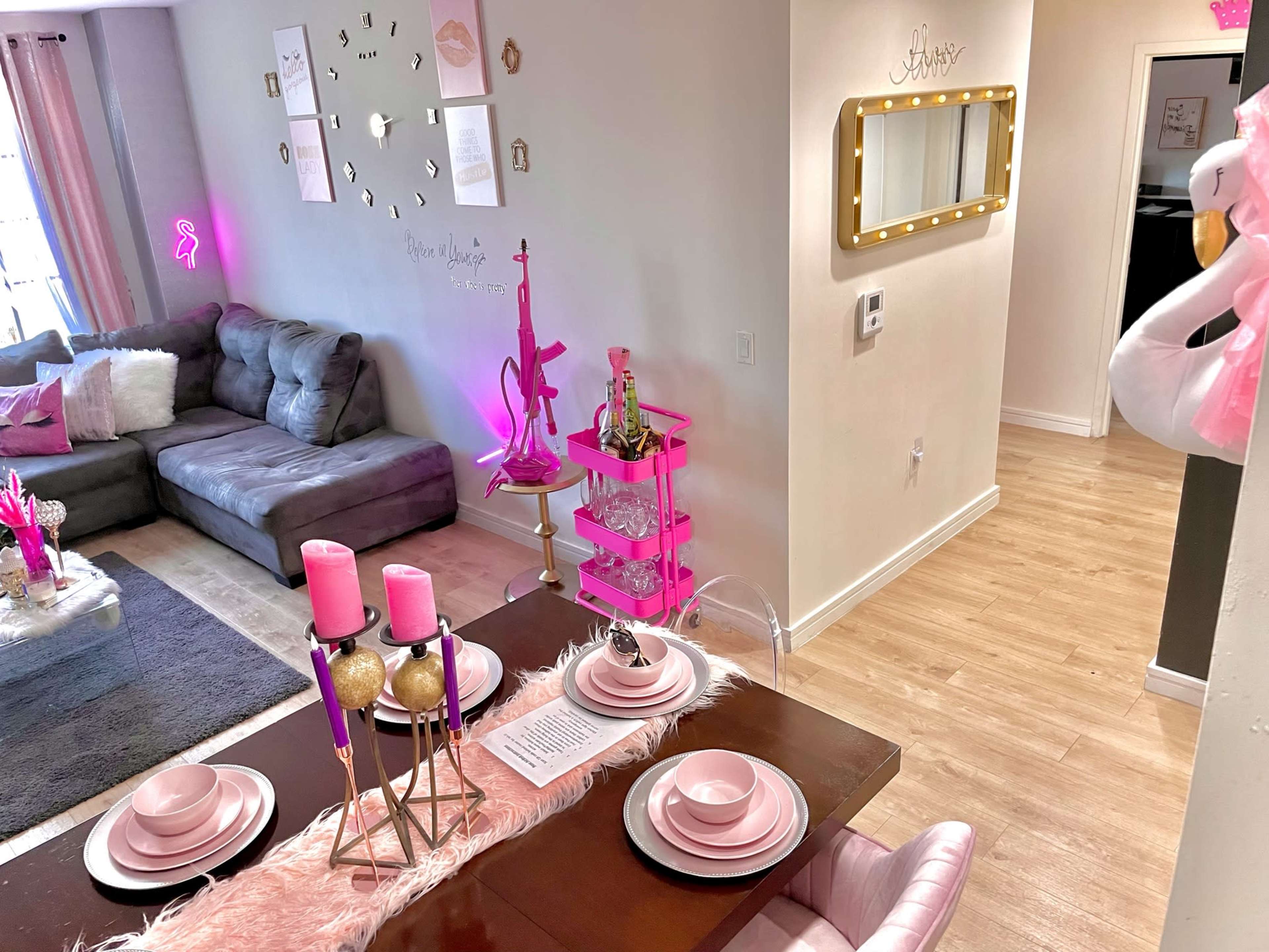 A dining area featuring a pink-themed table setup adjacent to a living room with a gray sofa and decorative elements.