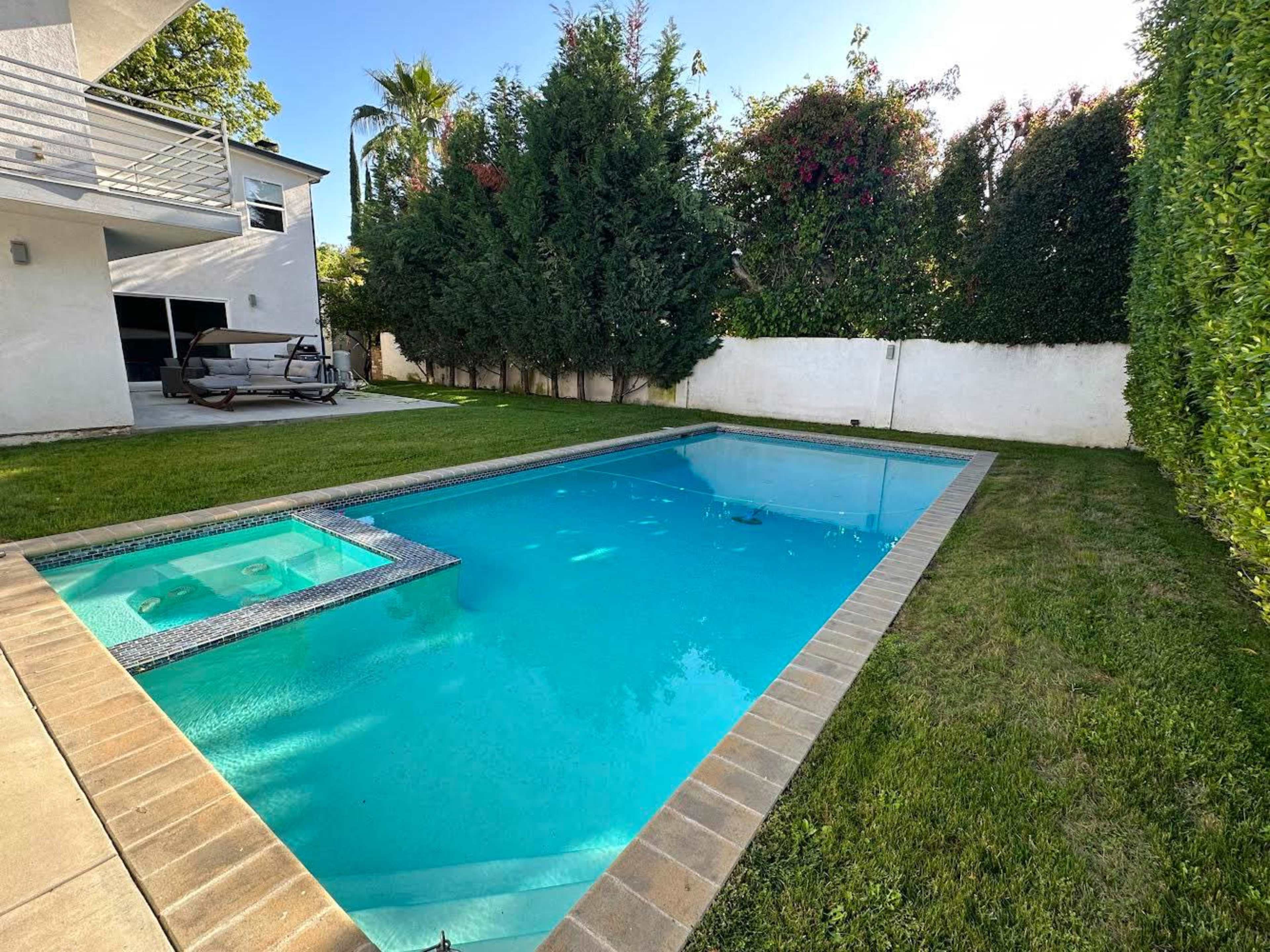 A clear swimming pool with a hot tub is situated in a grassy backyard surrounded by tall greenery and a white wall.