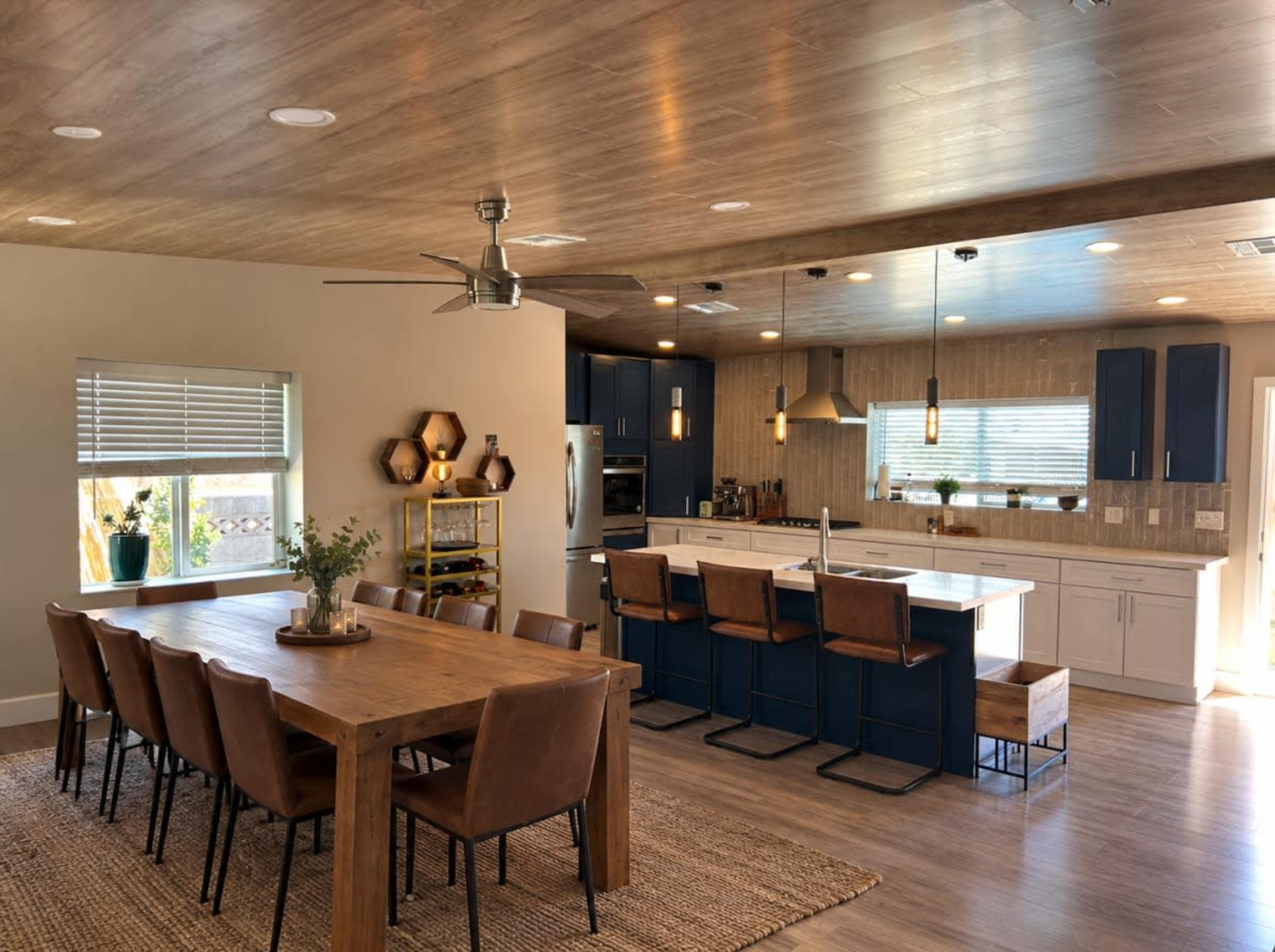 The image shows a modern kitchen and dining area with a large wooden dining table, leather chairs, and a kitchen island with bar stools, all under a wood-paneled ceiling.
