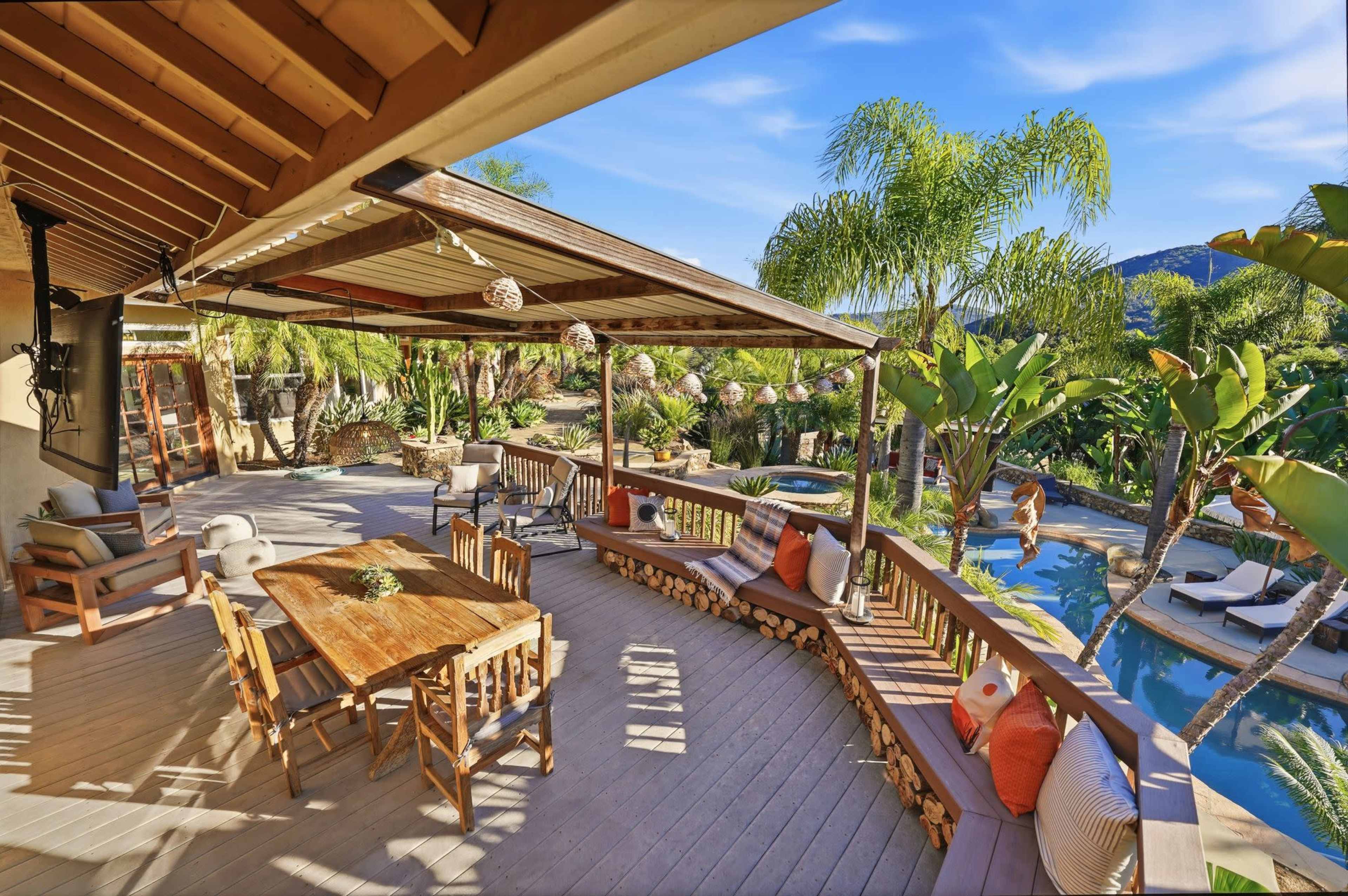 A wooden deck features a dining table and seating area, overlooking a swimming pool surrounded by lush greenery and palm trees.