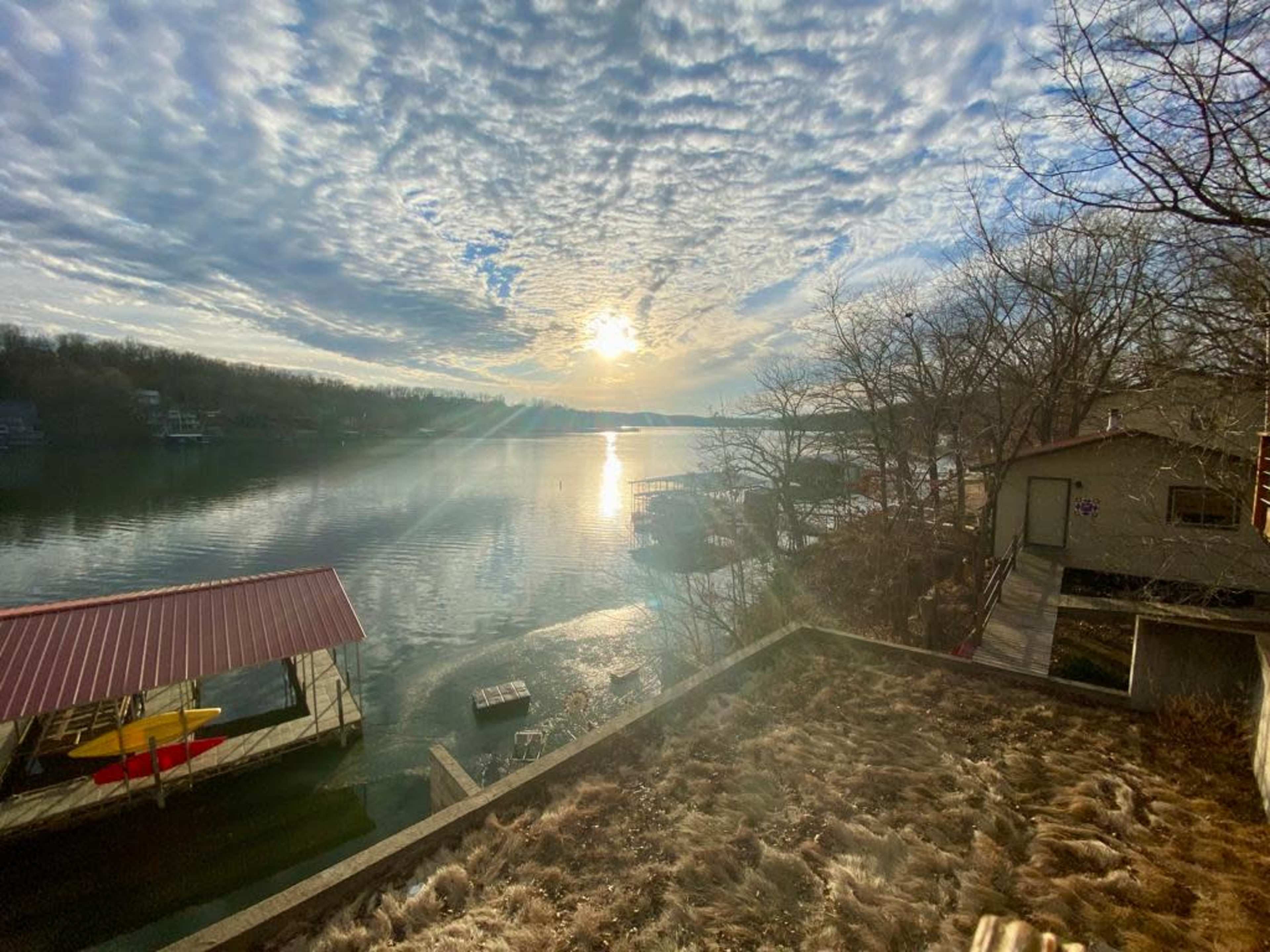 A serene lake scene with a setting sun reflecting on the water, framed by trees and a boathouse.