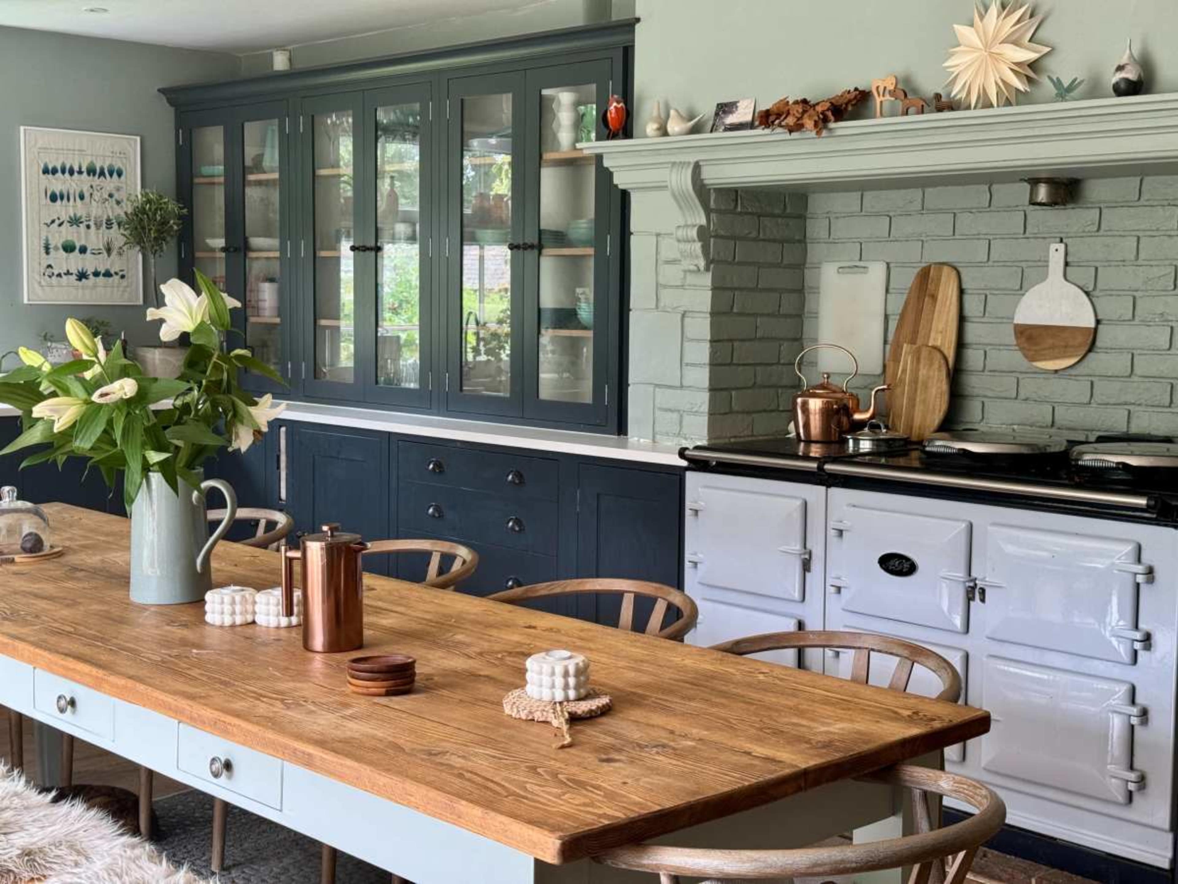 A modern kitchen features a wooden dining table surrounded by chairs, with a blue cabinetry backdrop and a stove integrated into a brick wall.