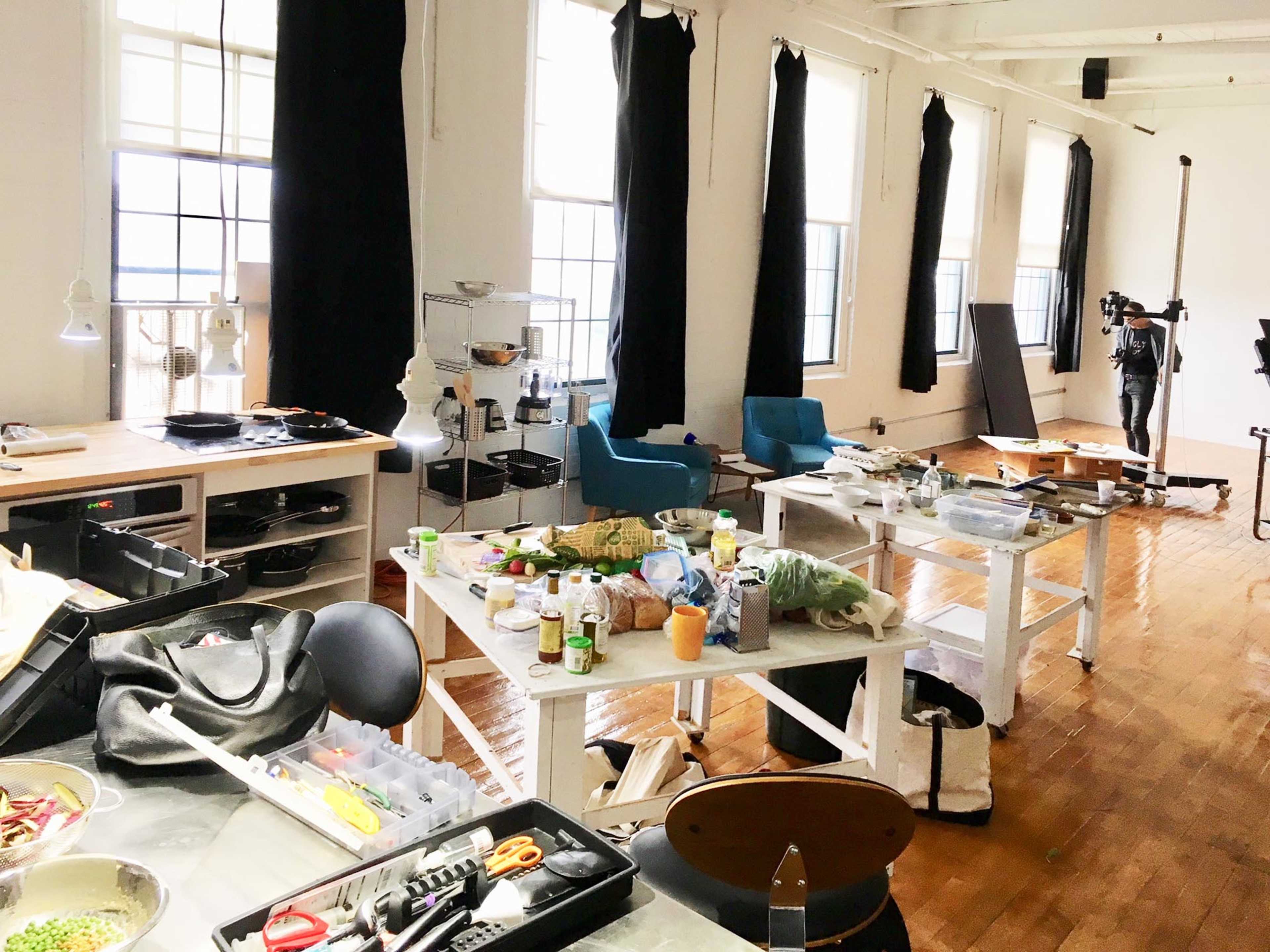 A spacious room with windows has several tables cluttered with kitchen items, utensils, and a few plants, alongside blue chairs and a mannequin in the background.