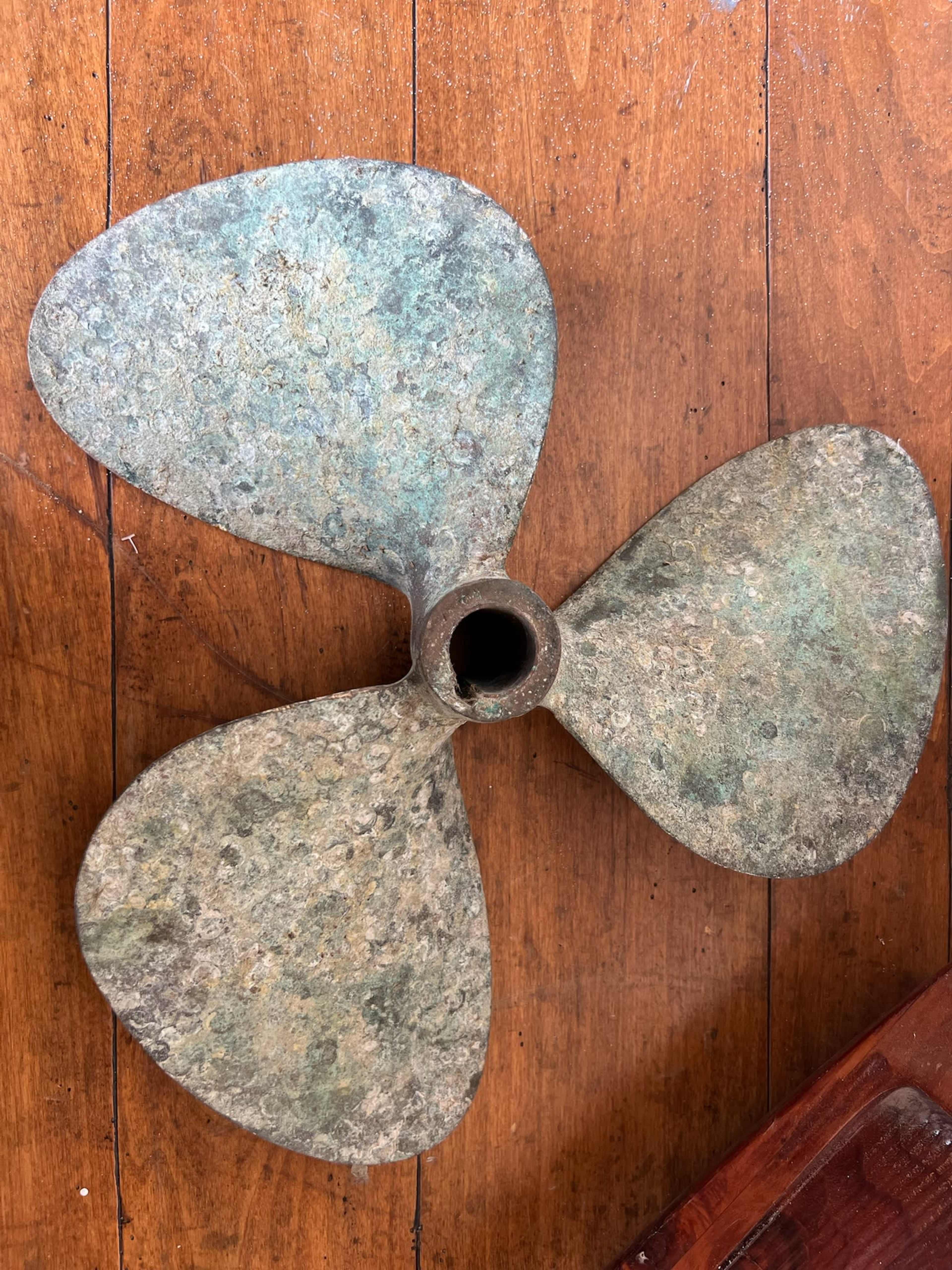 The image shows a three-blade bronze propeller resting on a wooden surface, showcasing its weathered and oxidized surface.