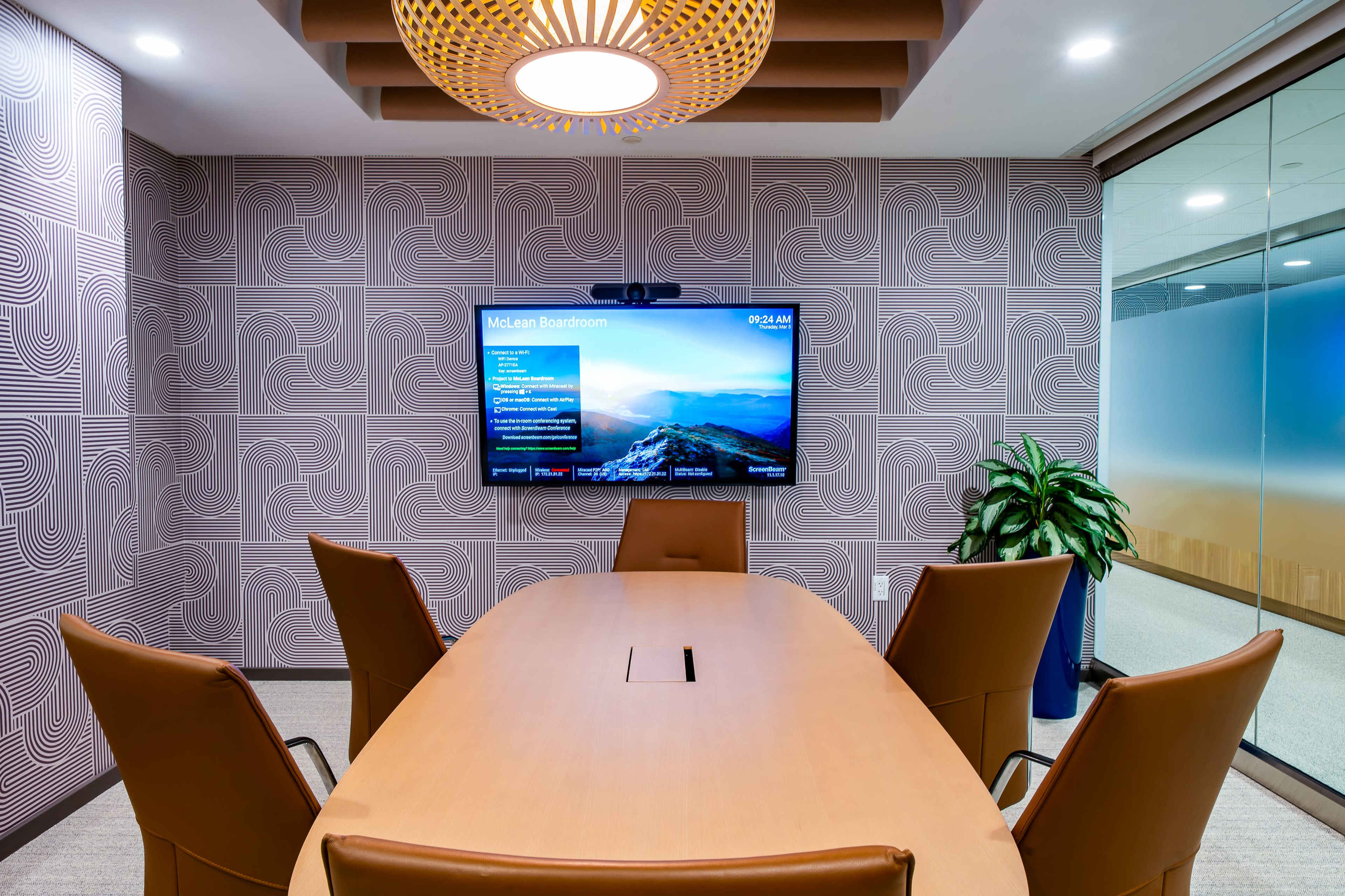 The image shows a modern conference room with a large wooden table, six brown leather chairs, a digital screen displaying information, and a plant in the corner.