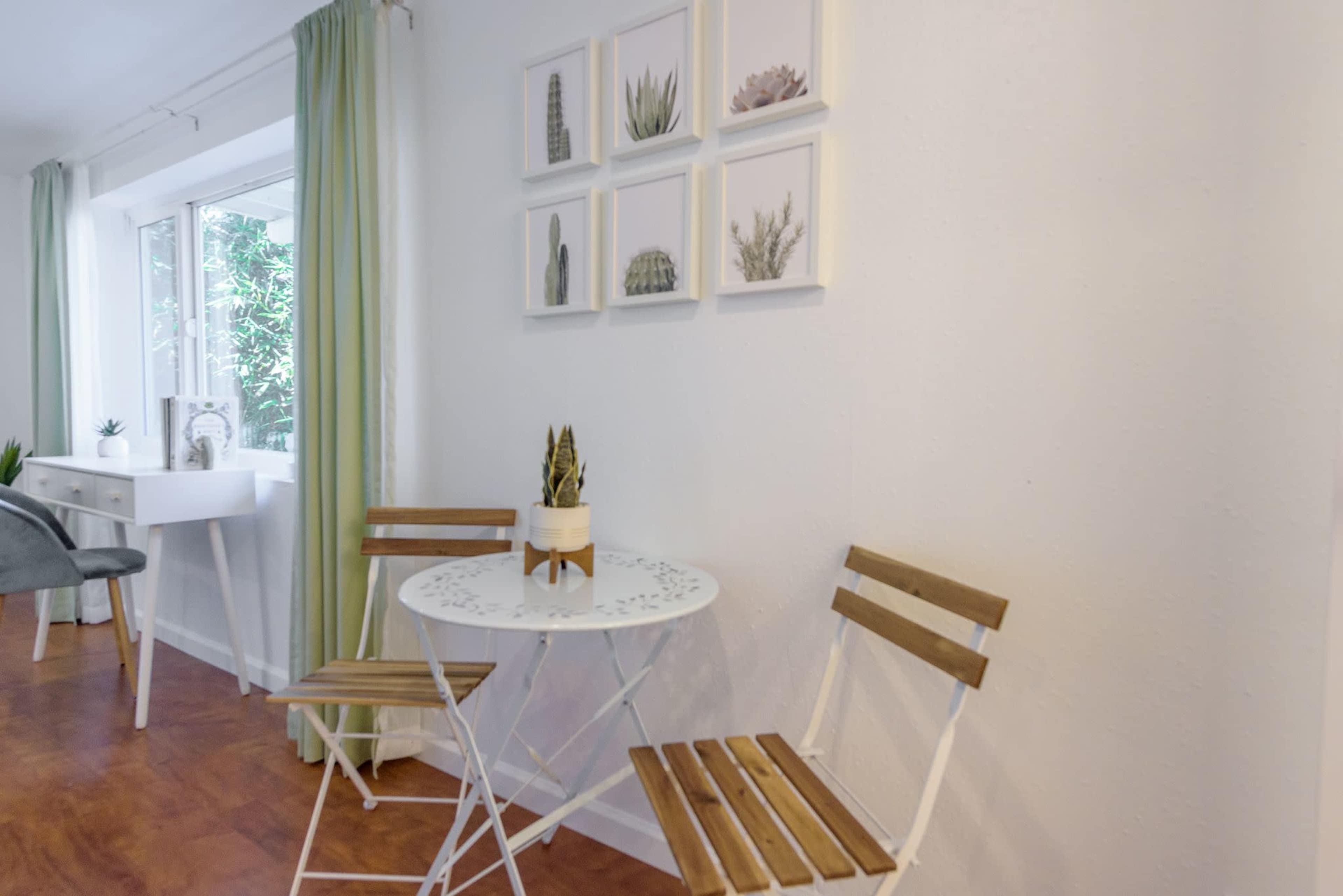A small dining area features a round white table with two wooden chairs, situated near a window adorned with light green curtains and framed cactus pictures on the wall.