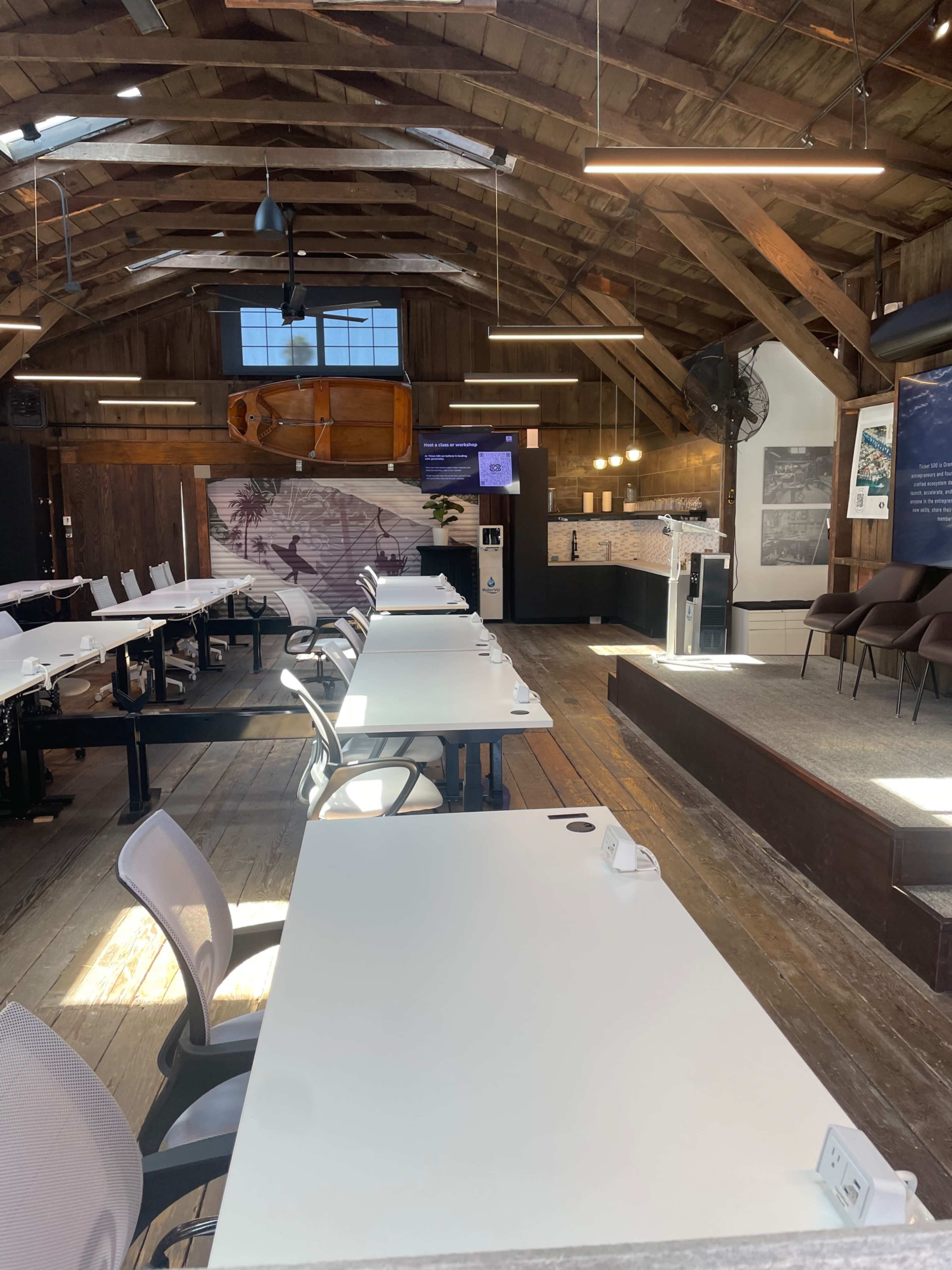 The image shows a spacious, well-lit meeting room with wooden beams, multiple tables arranged in rows, and a presentation screen at the front.
