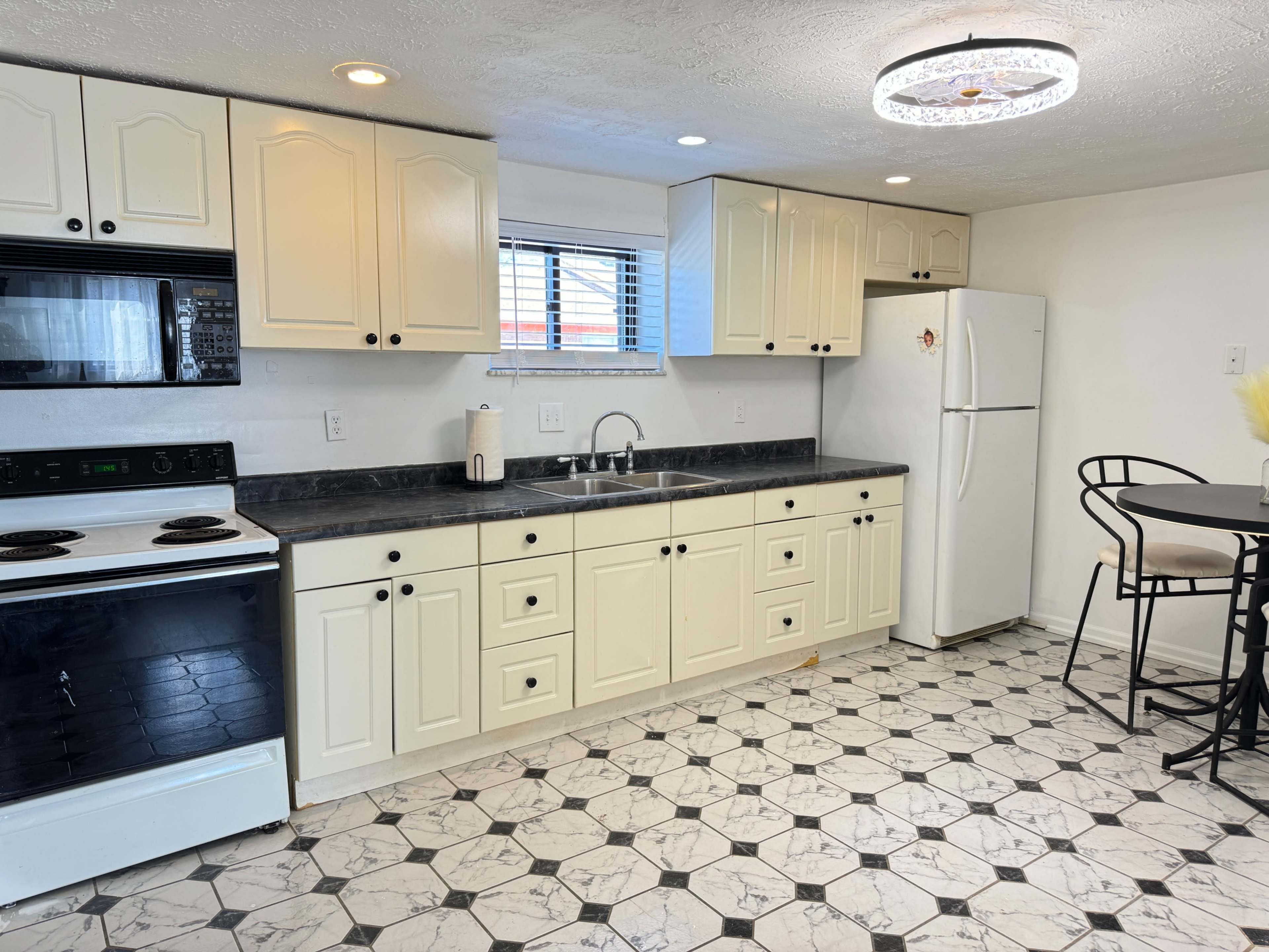 A kitchen with light-colored cabinets, a black countertop, a white refrigerator, an oven, and a small dining table with black chairs.