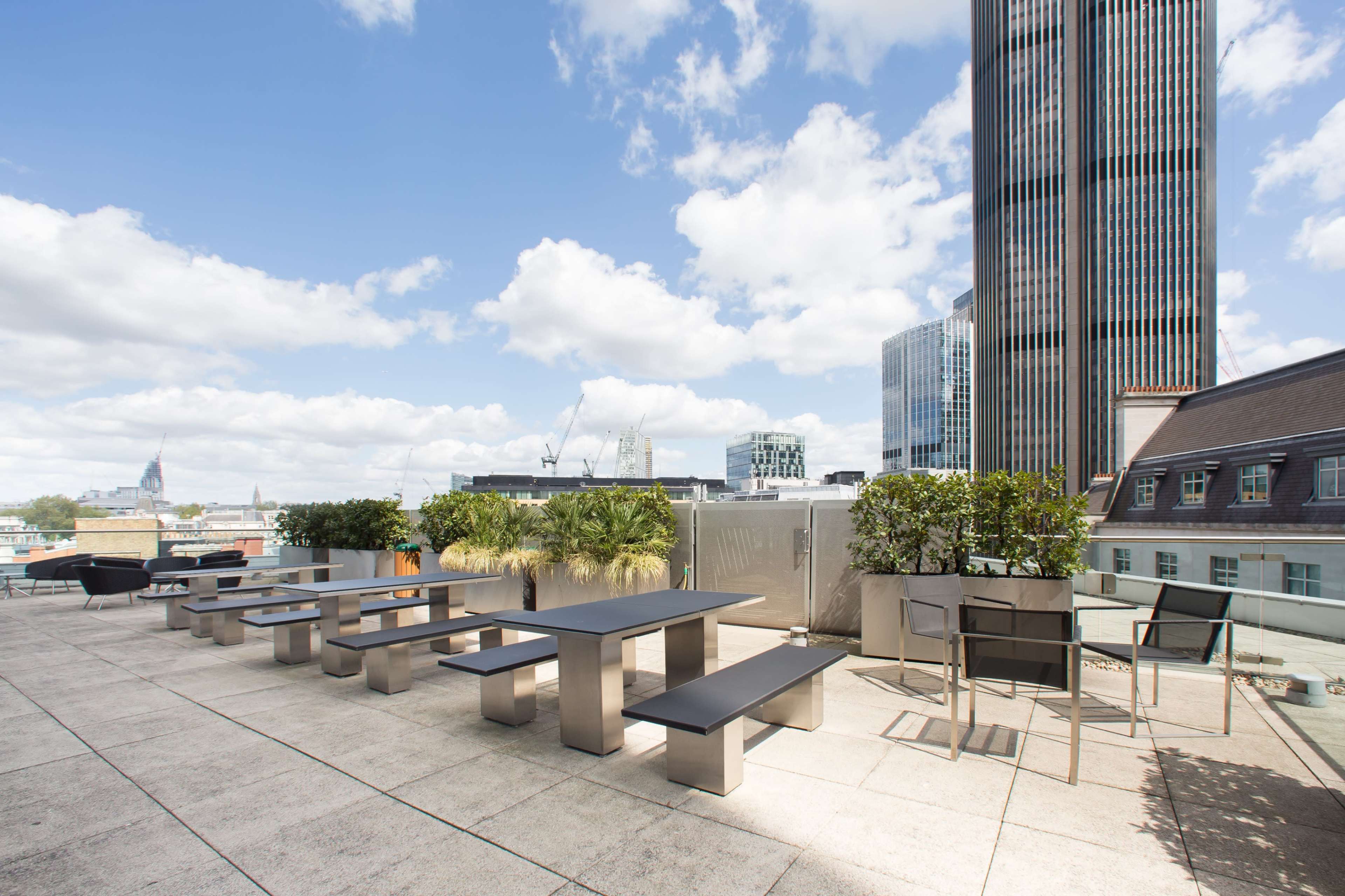 A rooftop terrace with modern tables and benches surrounded by potted plants, set against a backdrop of tall buildings and a blue sky.