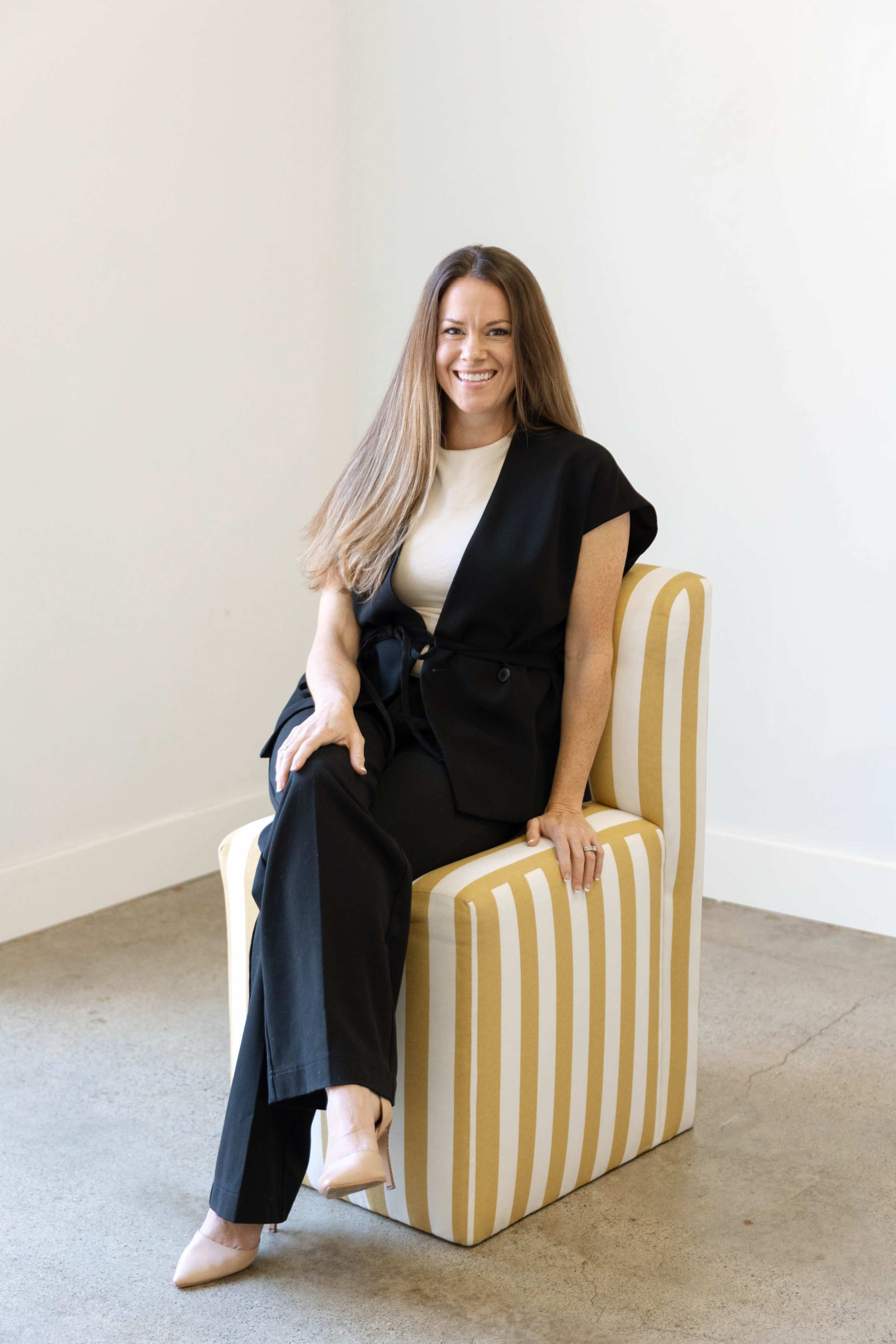 A person with long hair is sitting on a striped chair against a plain wall, wearing a black outfit with a white top.