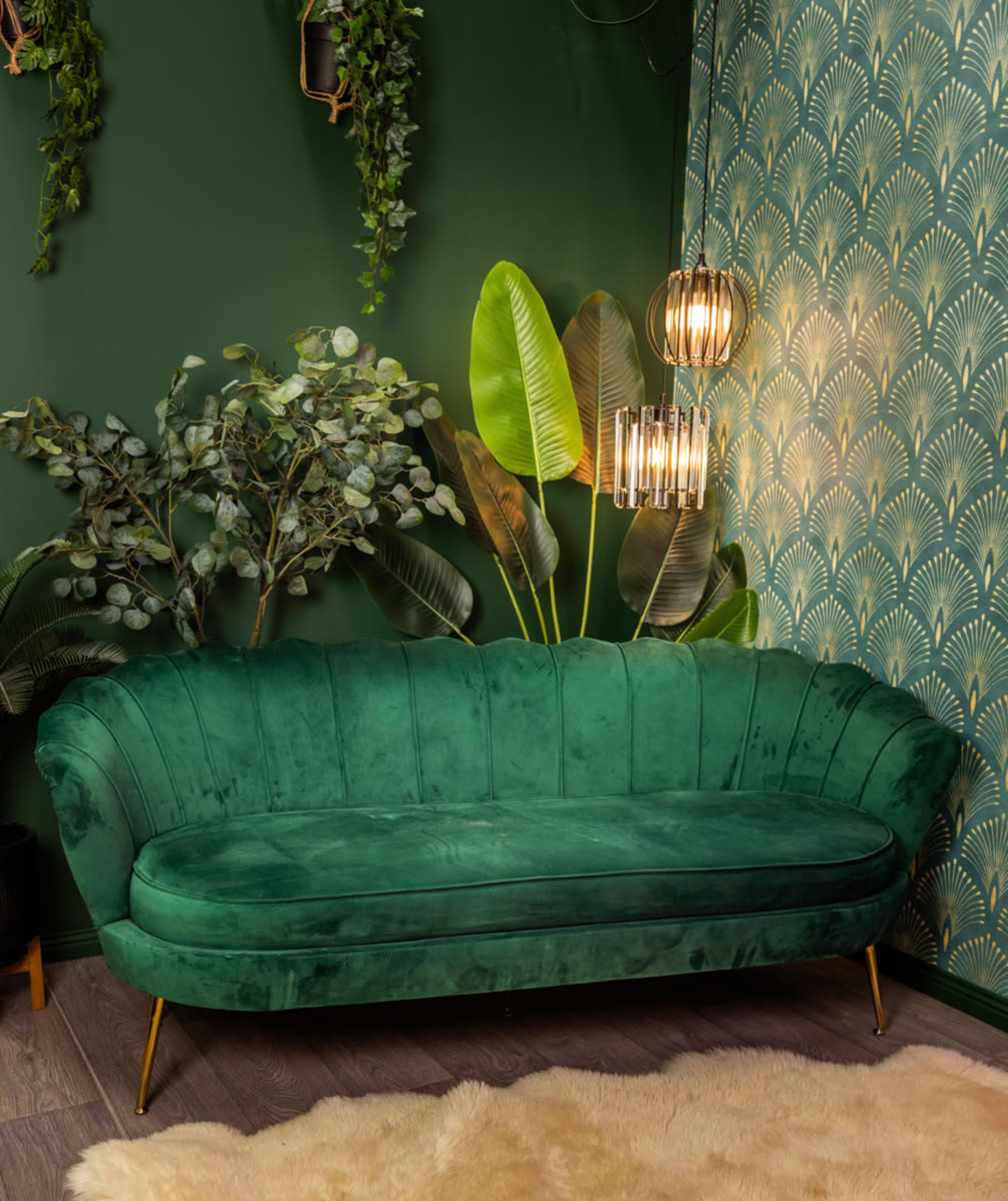 The image shows a green velvet curved sofa positioned against a dark green wall adorned with leafy plants and a patterned wallpaper featuring fan motifs, illuminated by pendant lights.
