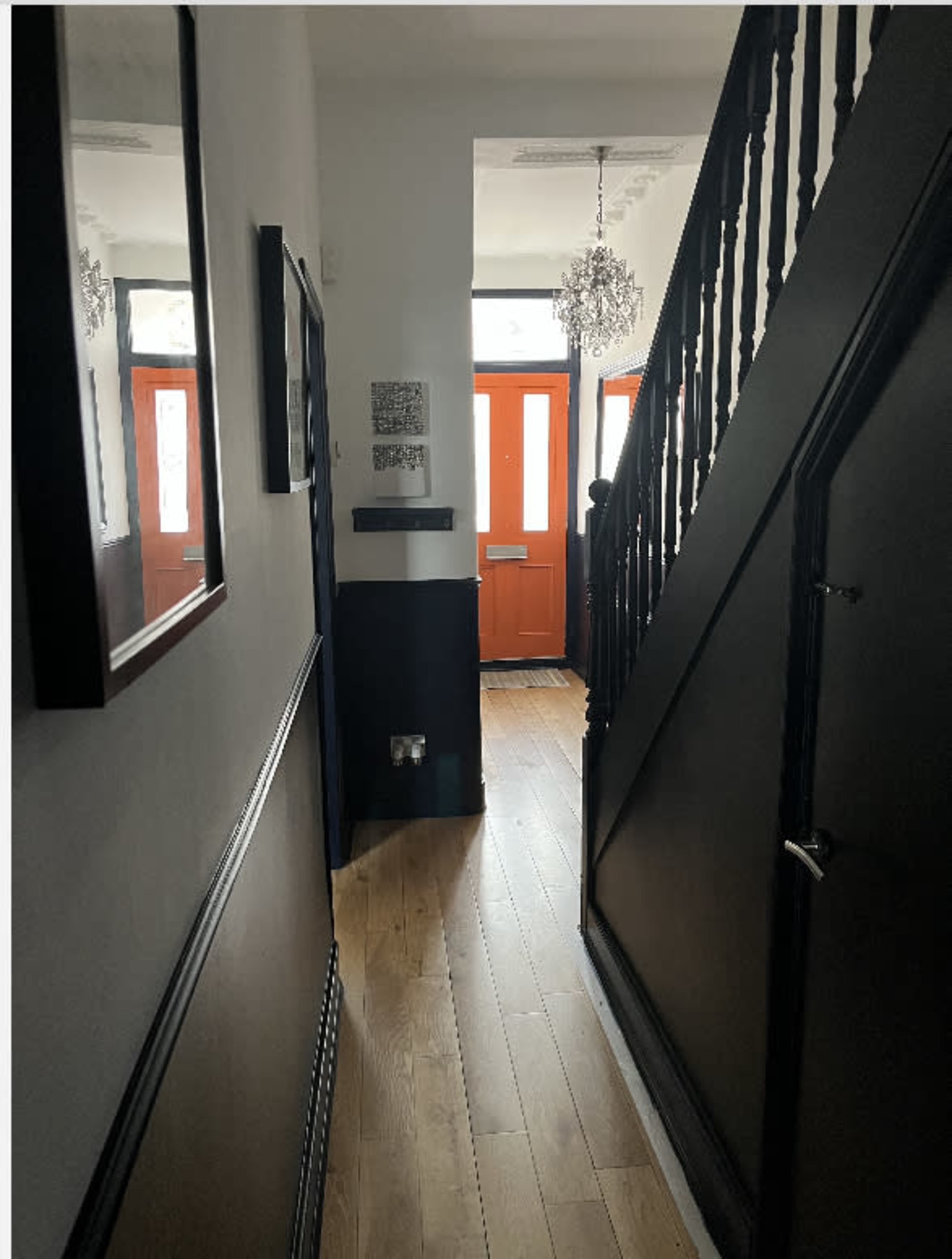 A hallway with wooden flooring, black walls, and a staircase leading up, illuminated by light coming from two orange doors at the end.