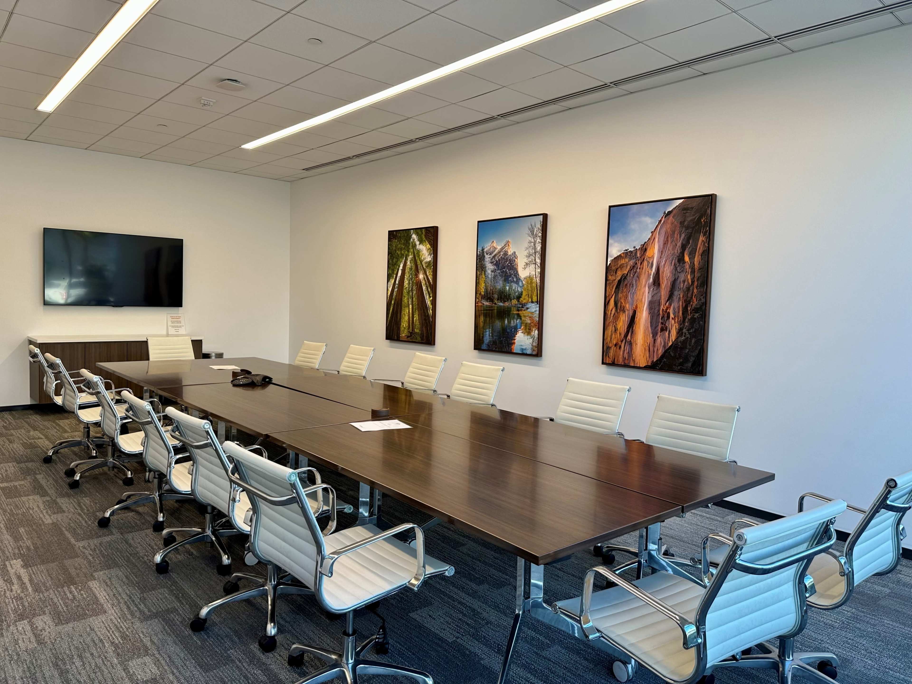 A large conference room features a long wooden table surrounded by white rolling chairs, with two framed landscape photographs on the walls and a television mounted on one side.