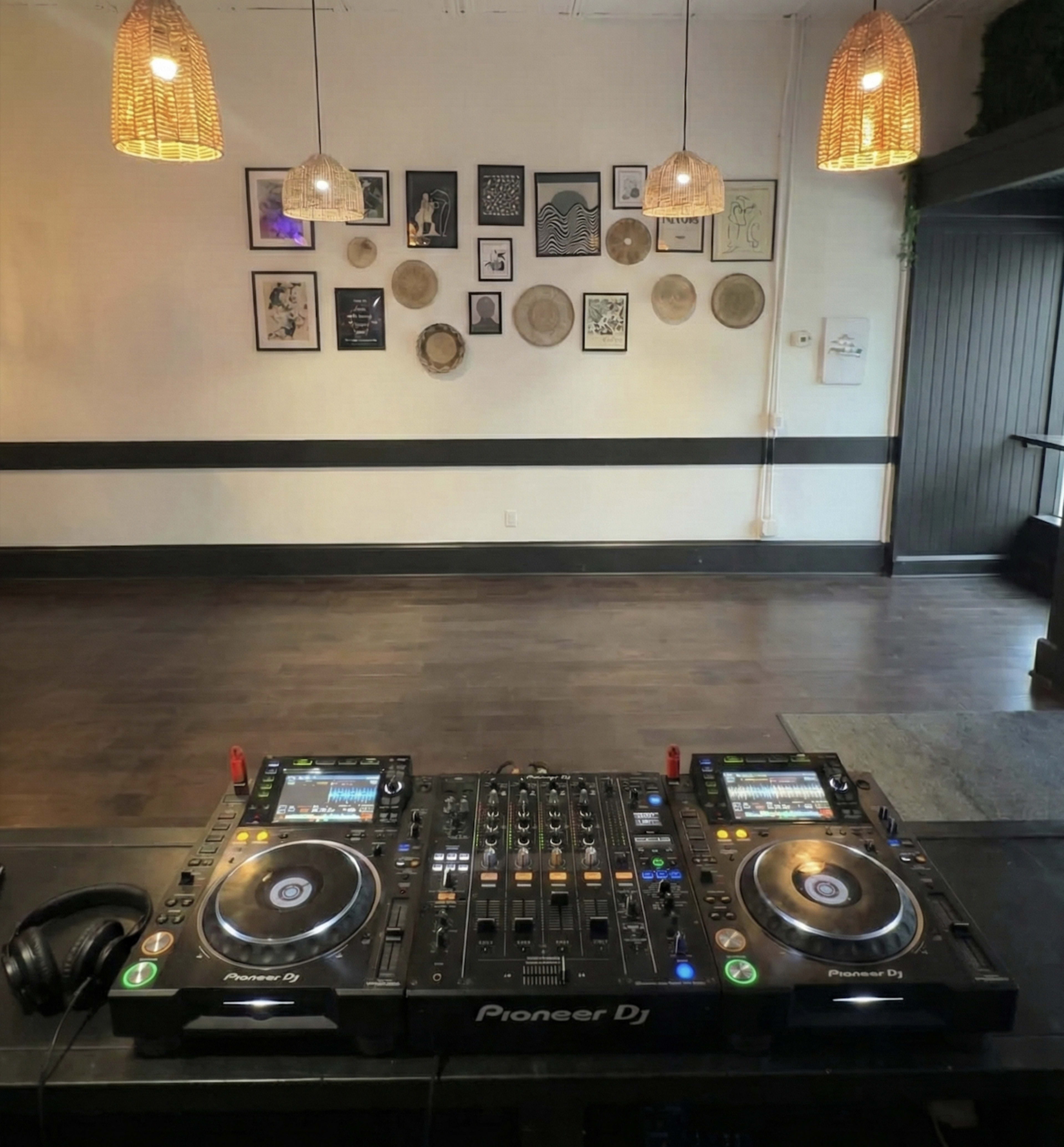 A DJ setup featuring two turntables and a mixer is positioned in the foreground, with a wall decorated with framed artwork and woven decor in the background.