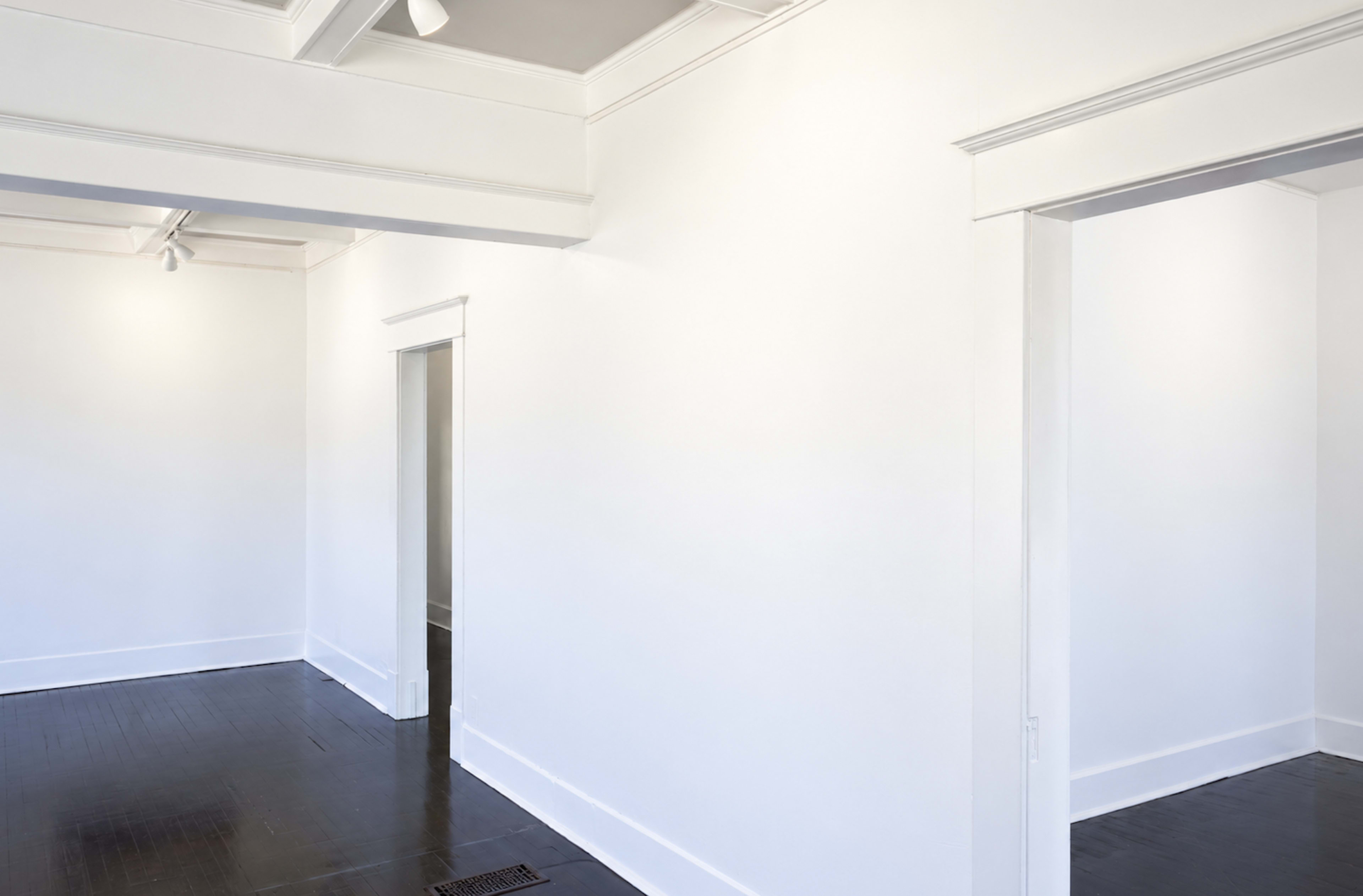 The image shows a spacious, empty room with white walls, hardwood floors, and an open doorway leading to another area.