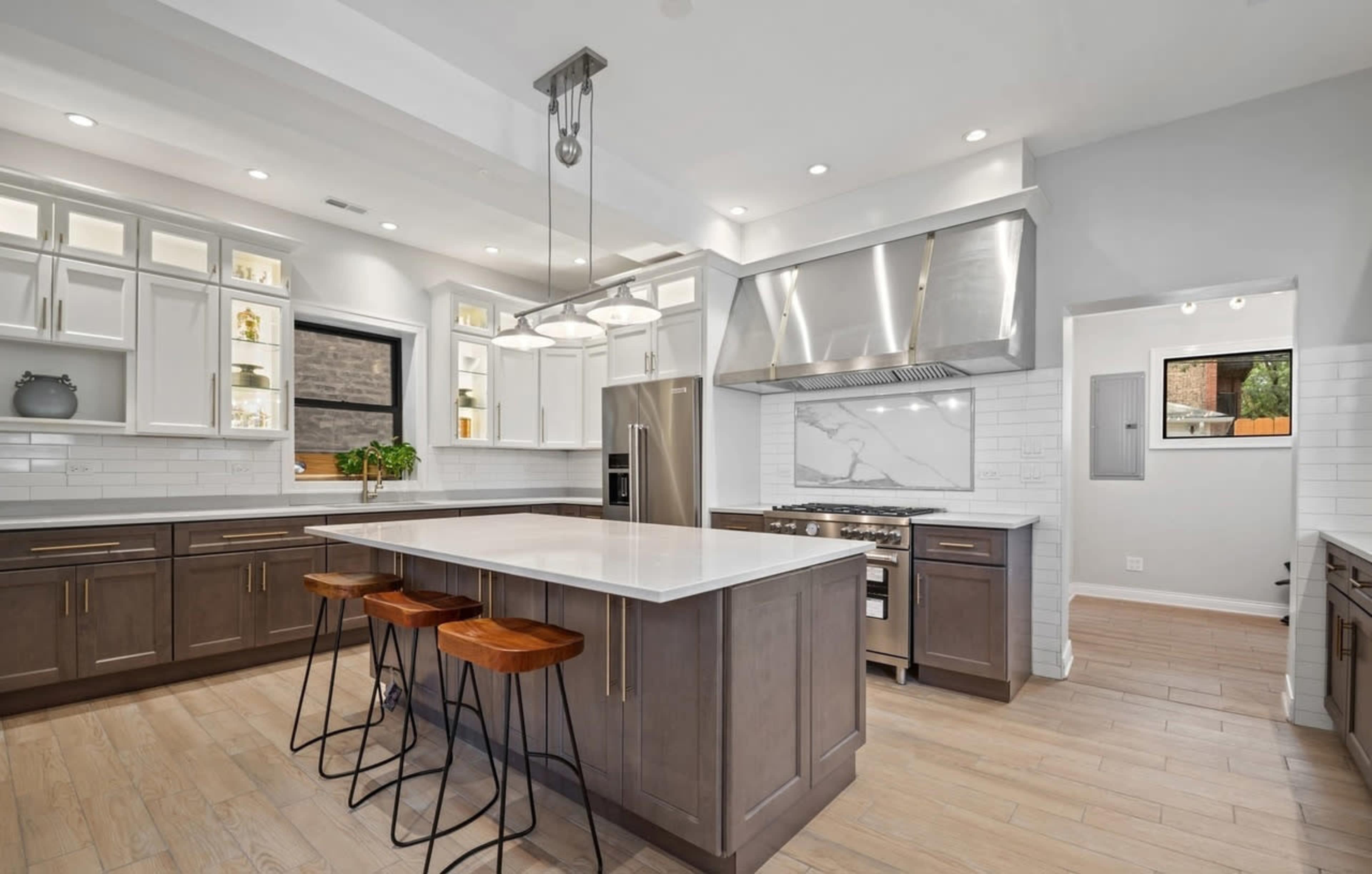 The image shows a modern kitchen featuring a large island with three bar stools, stainless steel appliances, and white cabinetry with a marble backsplash.