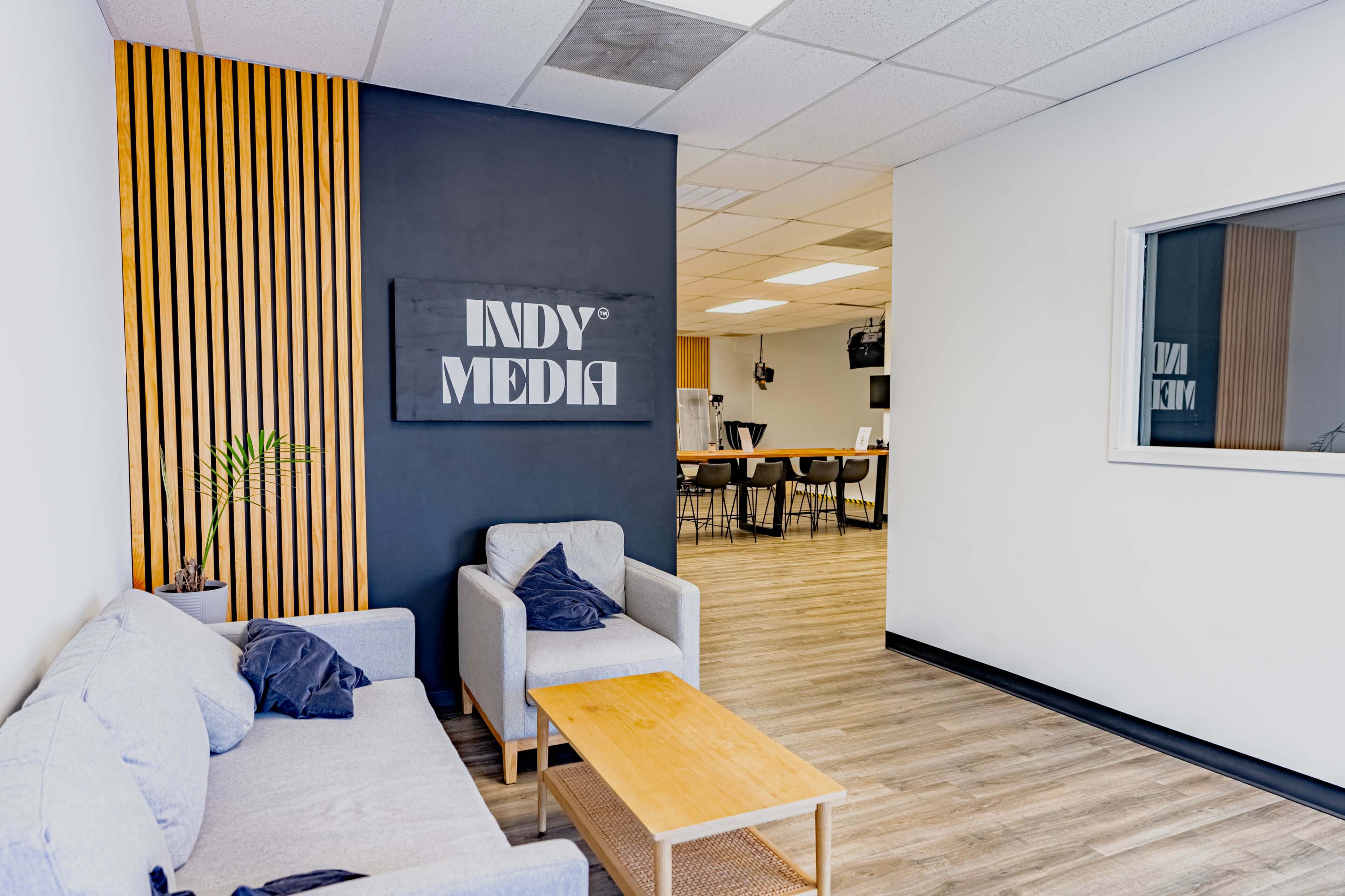 The image shows a modern office interior featuring a seating area with a gray sofa, coffee table, and a black wall displaying the sign "INDY MEDIA," along with a view of a workspace in the background.