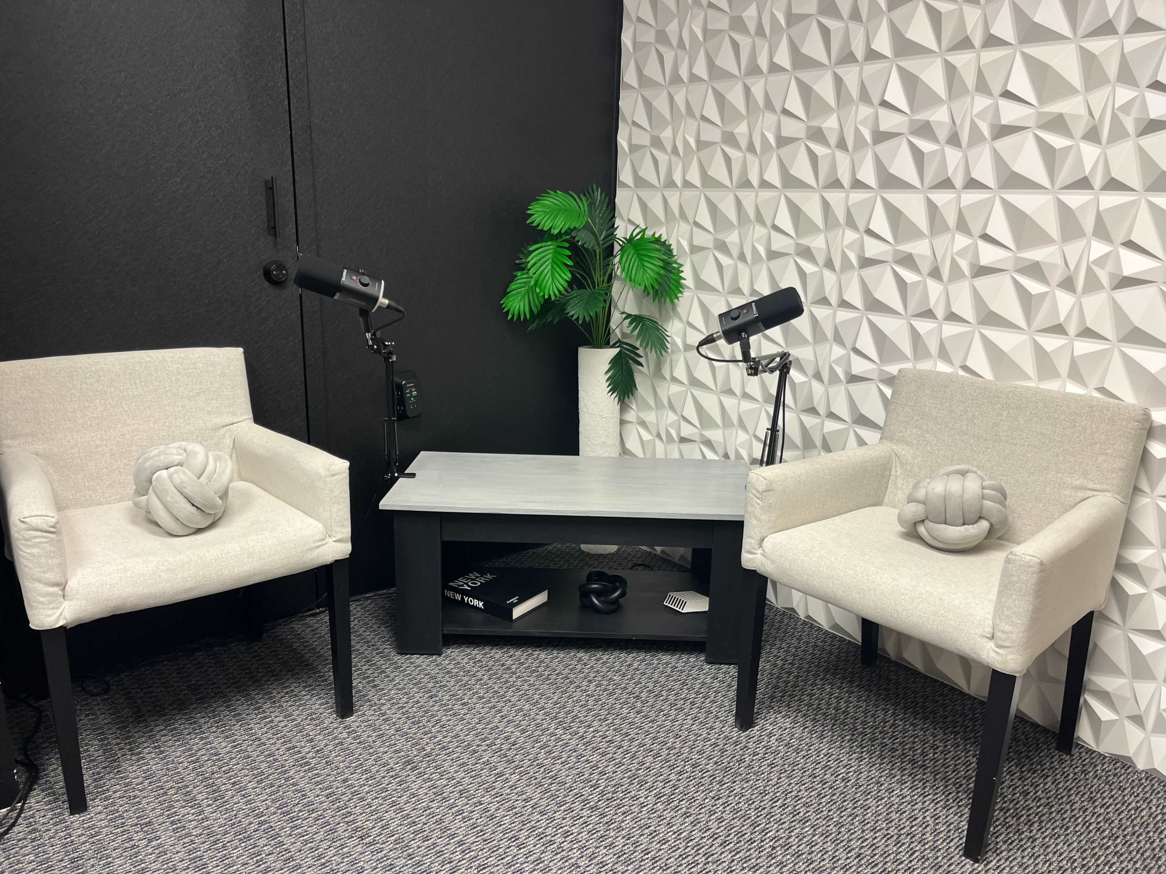 The image shows a small, modern room with two light-colored chairs facing a black coffee table, surrounded by a textured white wall and a decorative plant.