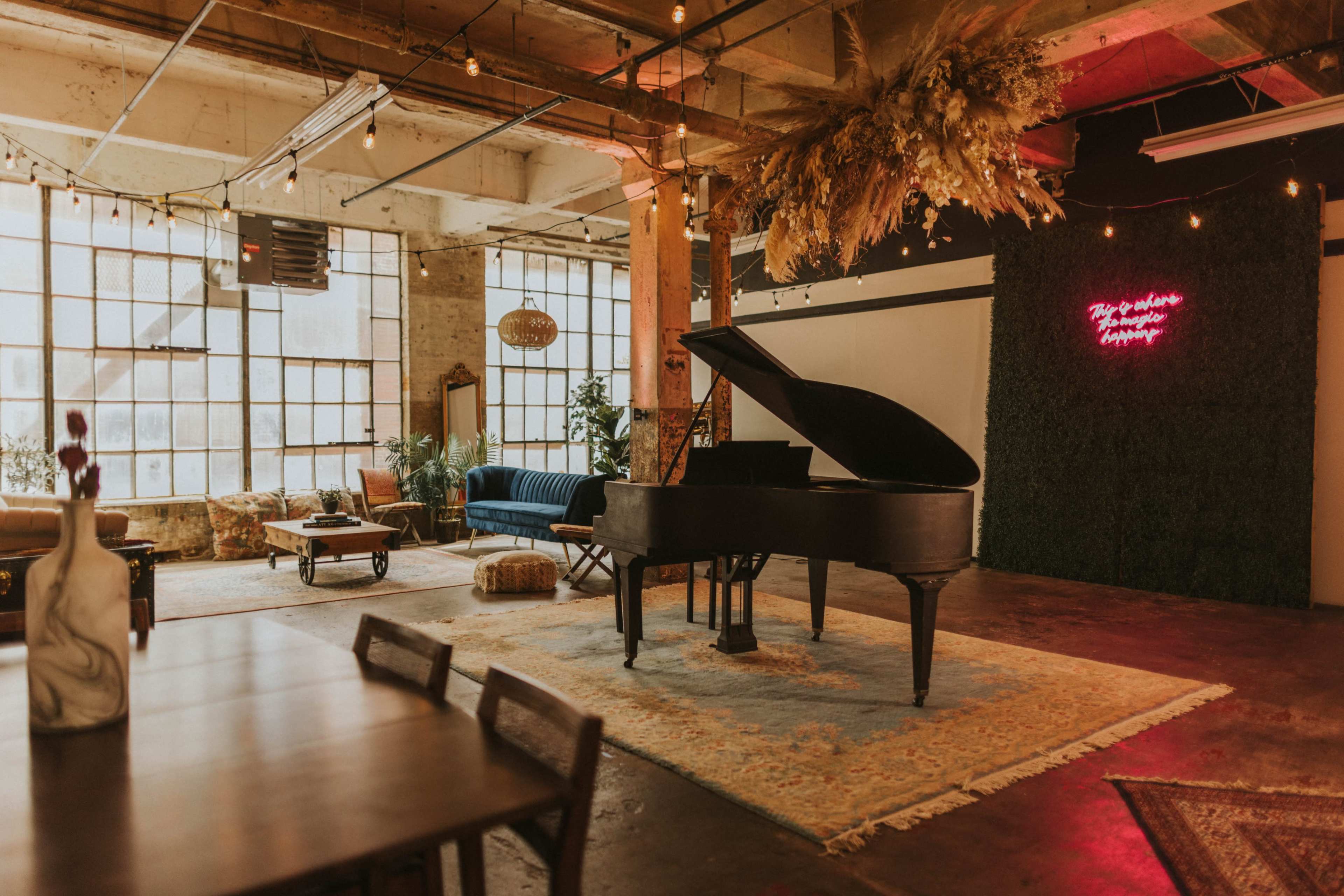 A grand piano sits on a rug in a spacious, well-lit room with large windows, surrounded by modern seating and decorative plants.