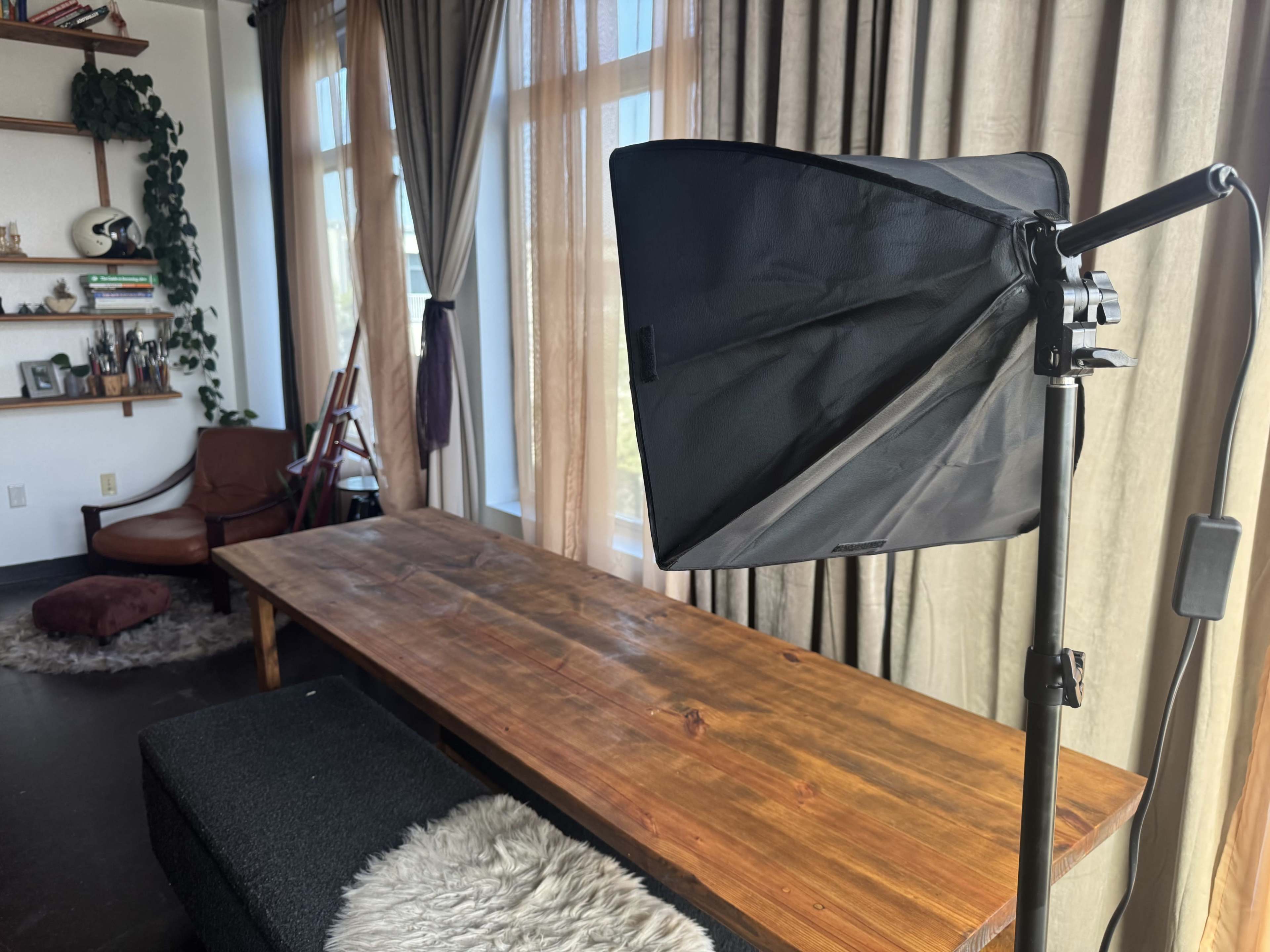 A black softbox light is positioned next to a wooden table in a room with large windows and beige curtains.