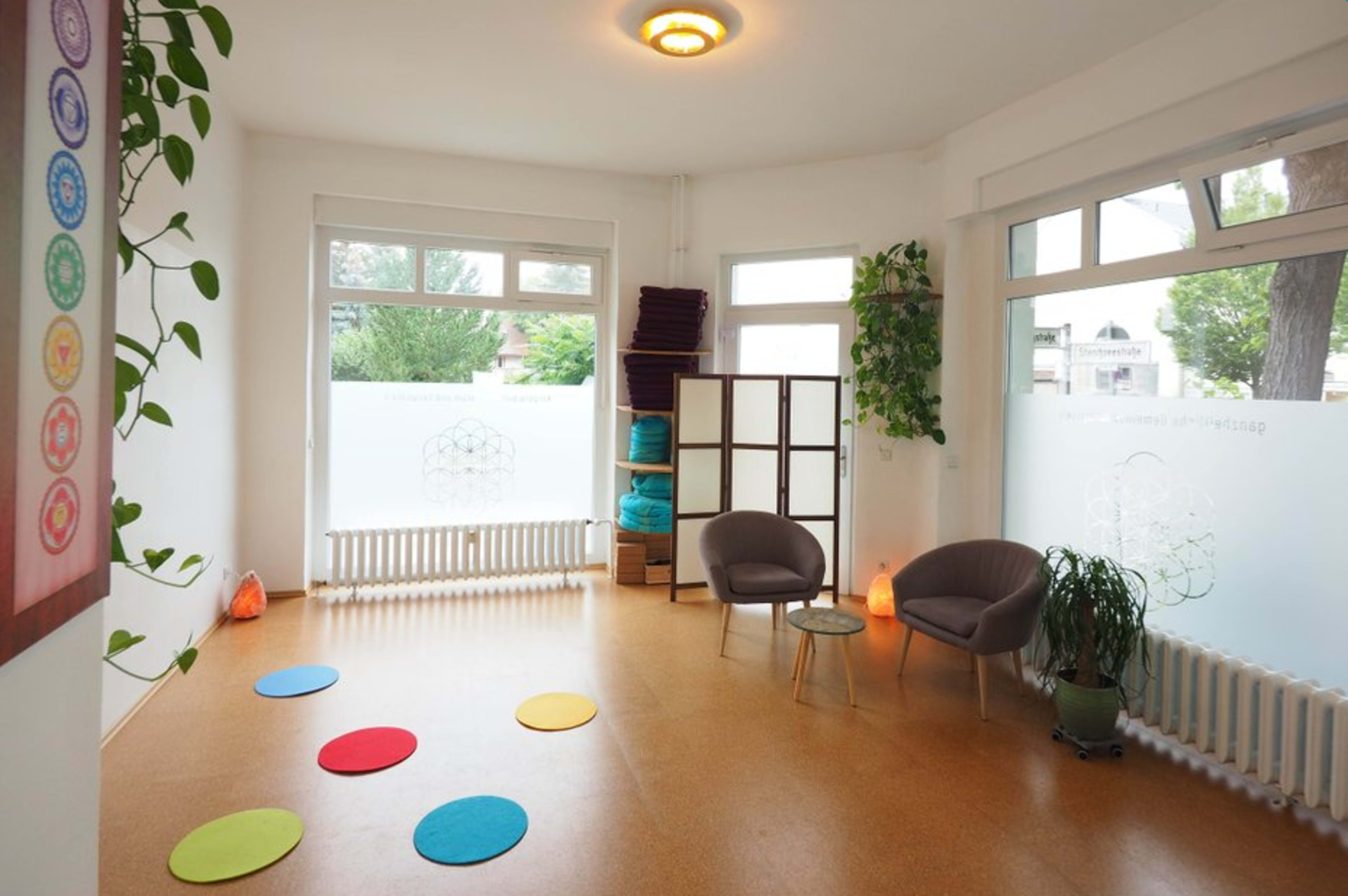 A bright, minimalist room with two chairs, colorful circular rugs on the floor, plants, and large windows allowing natural light.