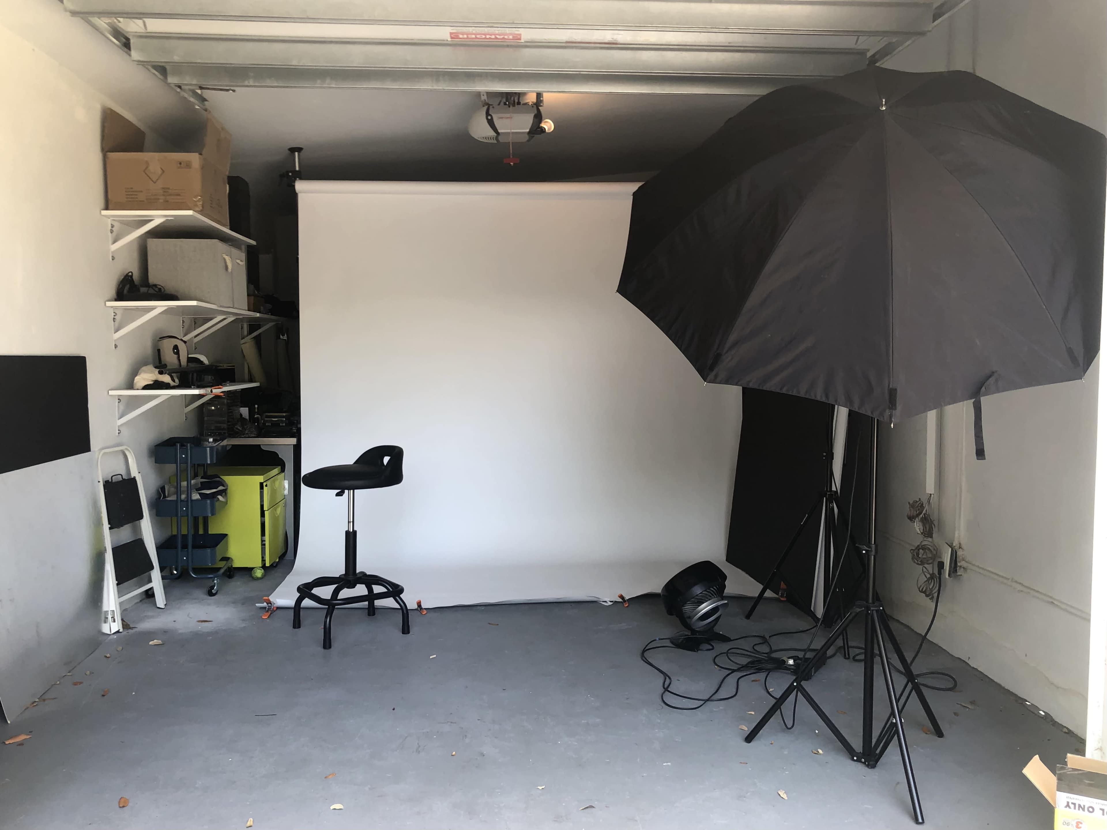 The image shows a garage setup with a white backdrop, a black umbrella light, a stool, and various equipment on shelves.