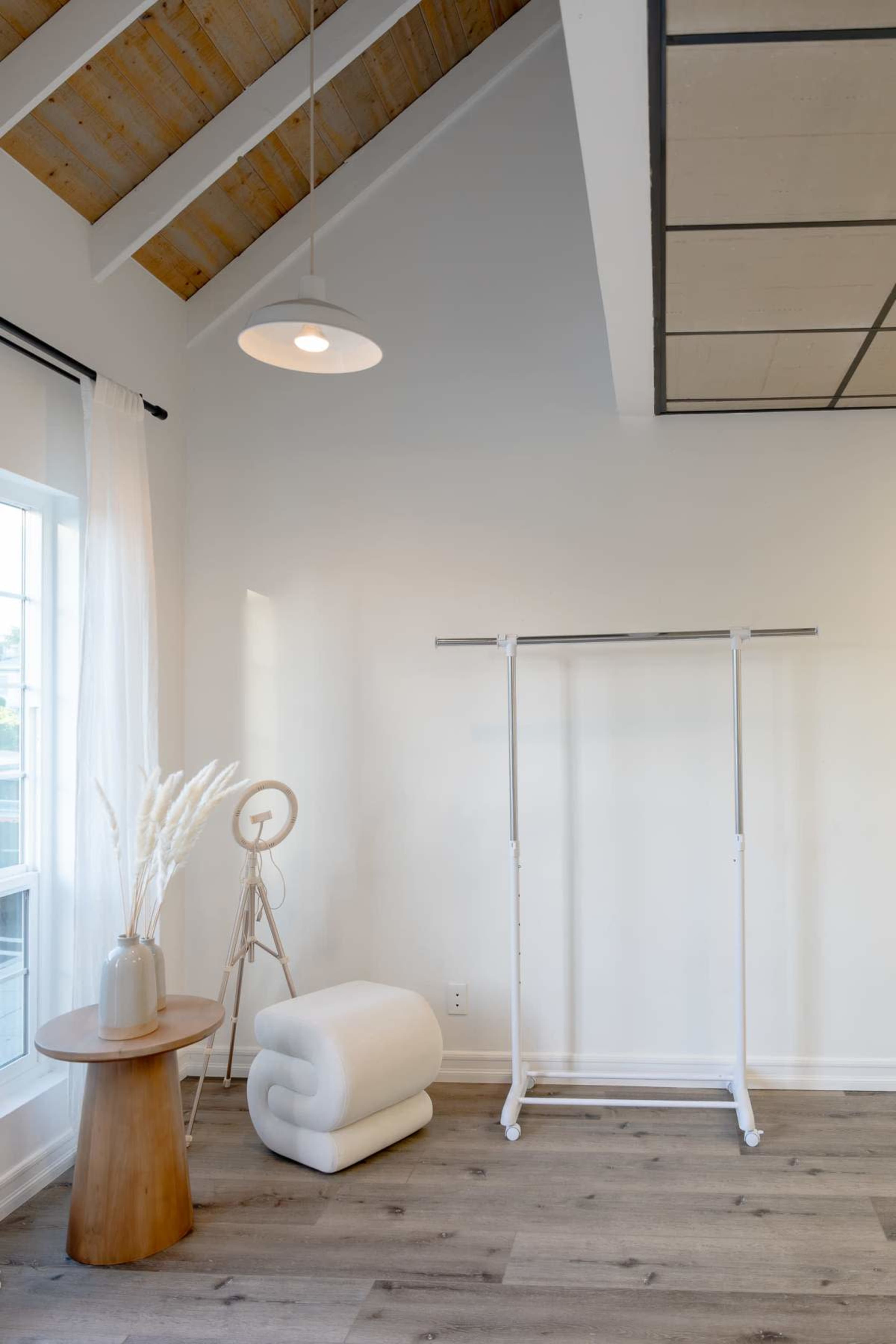 The scene features a minimalistic interior with a clothing rack, a rolled white blanket, and a table with a vase on a wooden floor beneath a pitched ceiling.