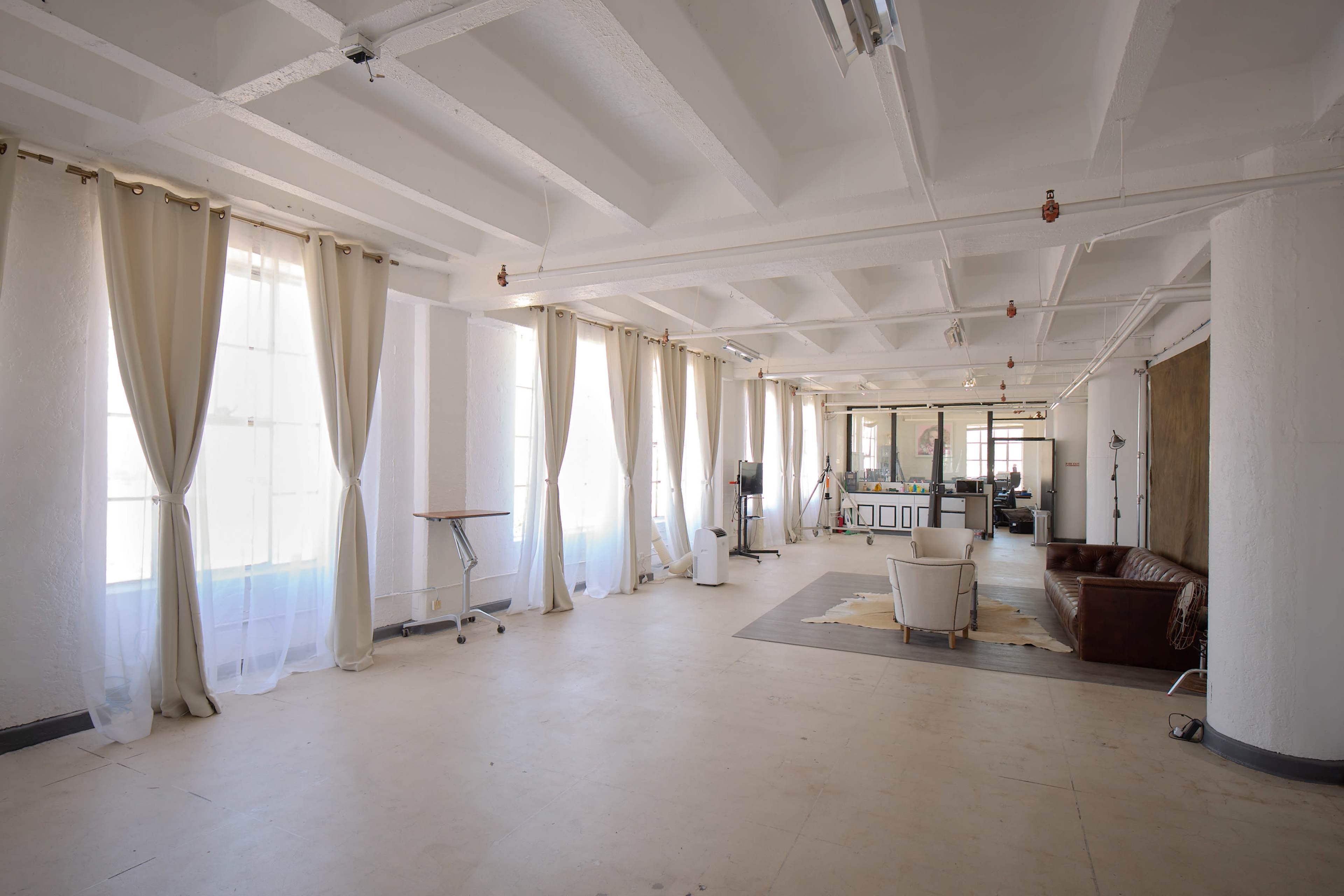A spacious, empty room with large windows, sheer curtains, and minimal furniture, including a white chair and a brown couch.