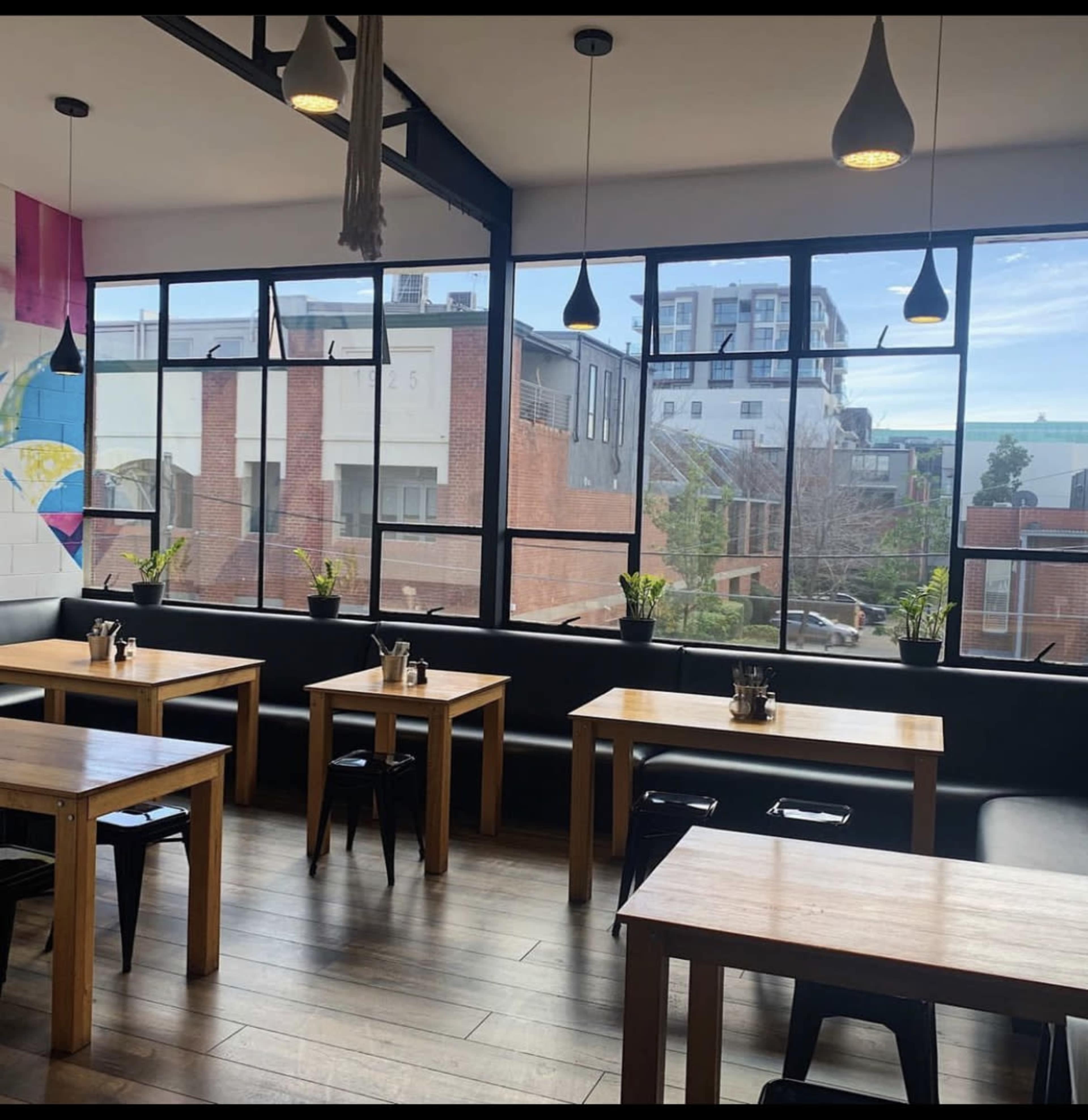 A modern restaurant interior with wooden tables, black chairs, and large windows displaying an urban view.