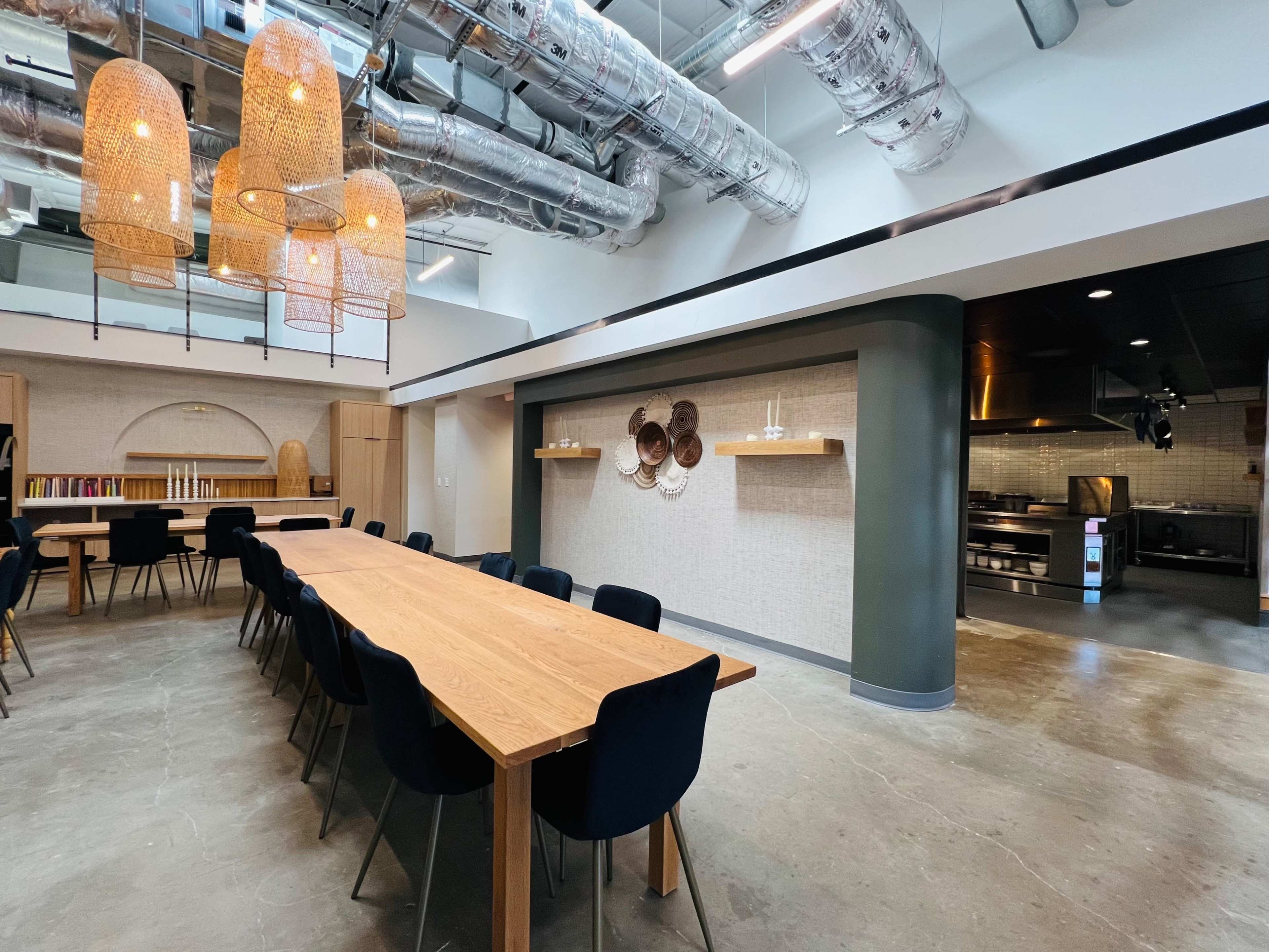 A spacious dining area features a long wooden table surrounded by black chairs, with high ceilings and industrial-style light fixtures above.