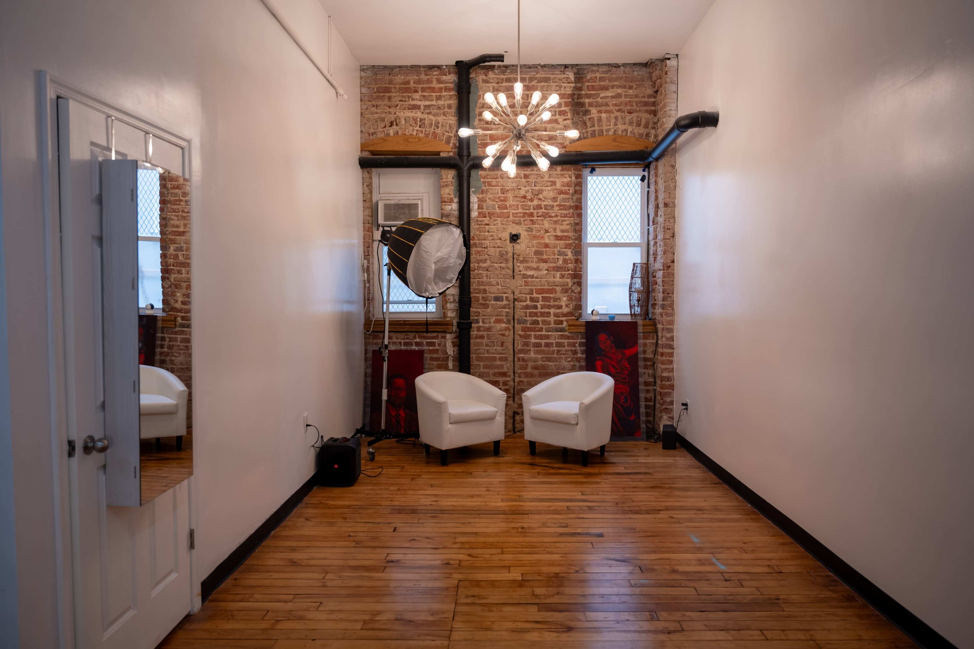 The image shows a minimalist room with exposed brick walls, featuring two white chairs, a large softbox light, and a modern chandelier.