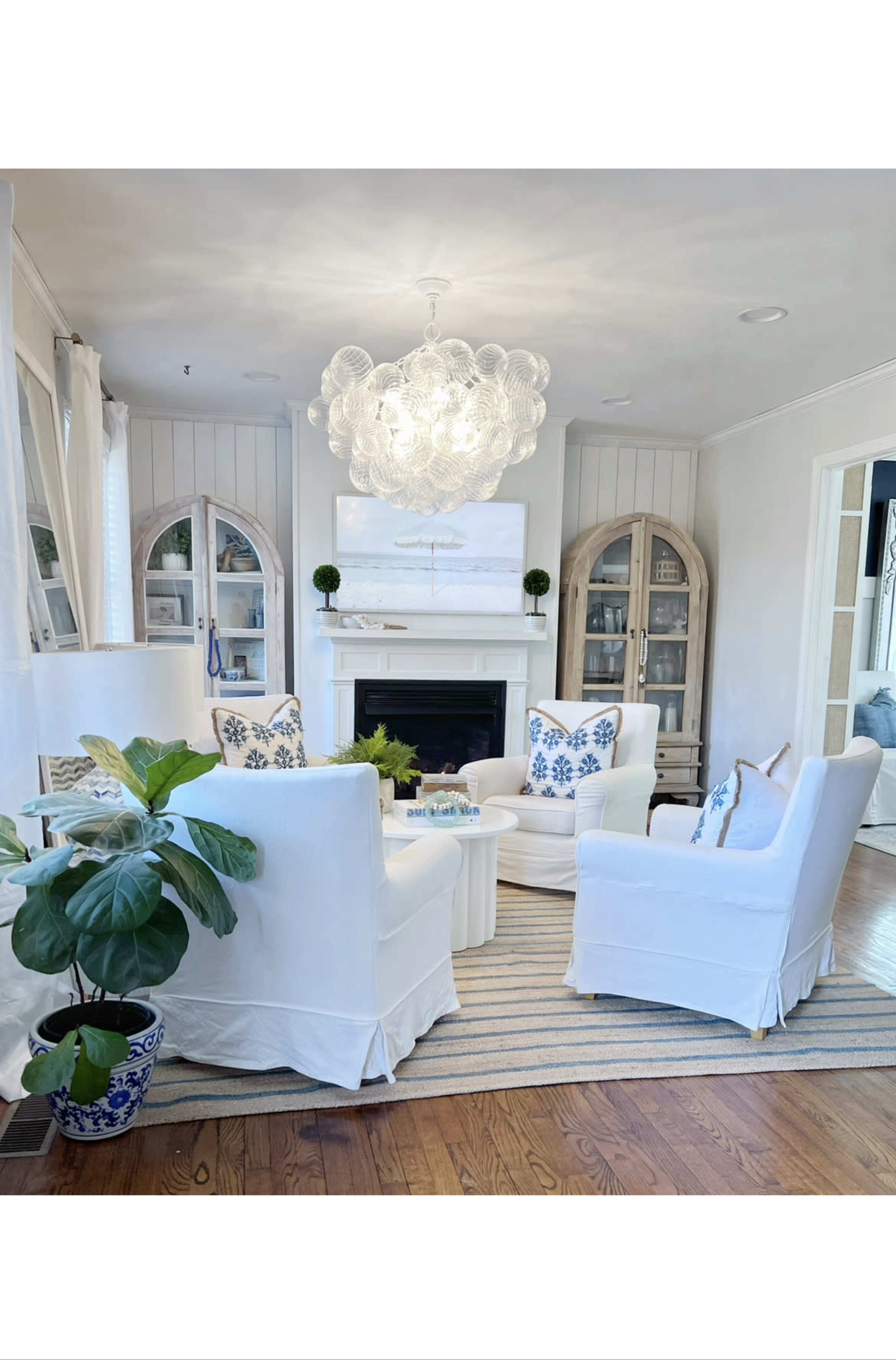 A cozy living room features white upholstered chairs around a white coffee table, with a large chandelier overhead and a fireplace in the background.