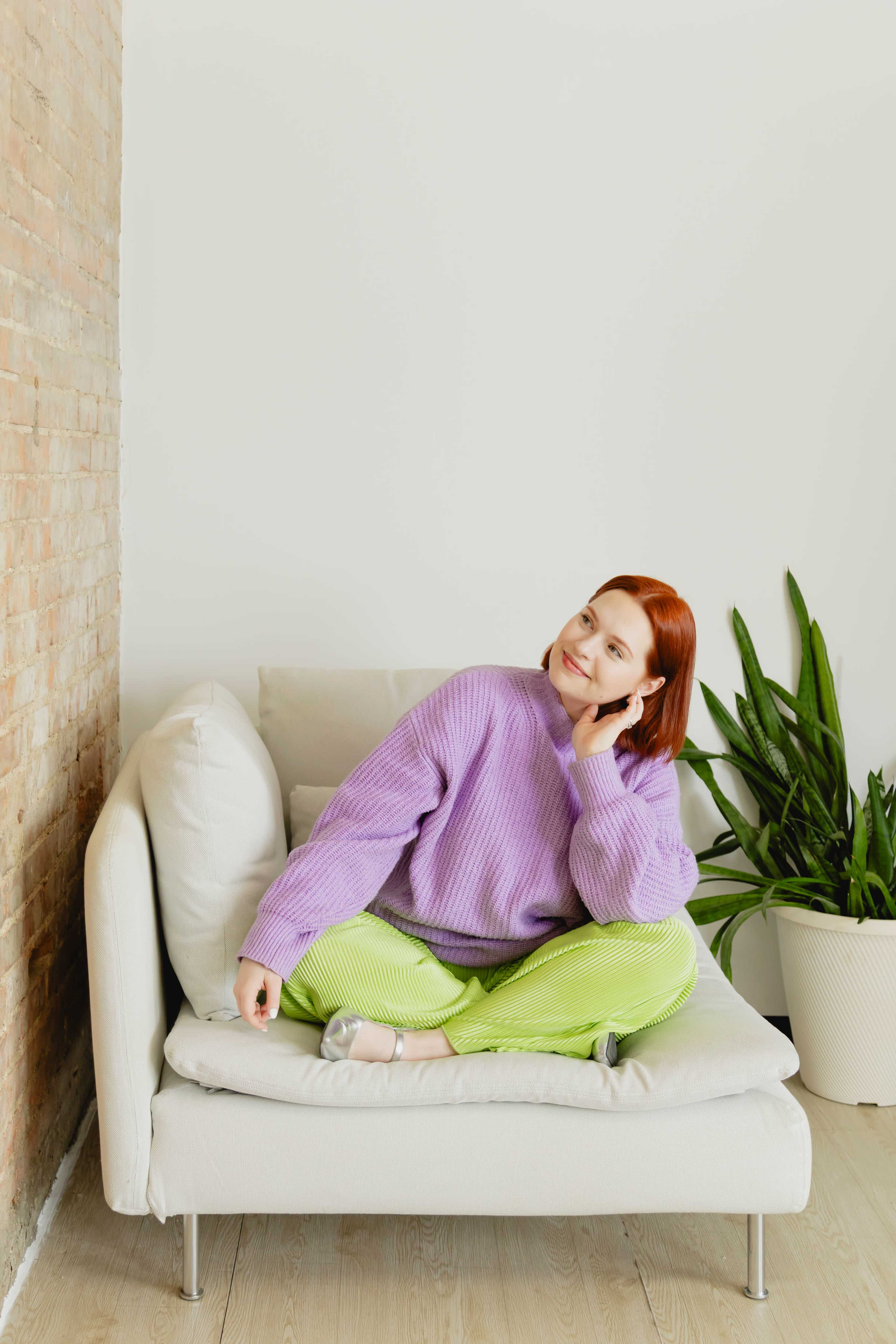 A woman with red hair sits cross-legged on a light-colored sofa in a bright room next to a potted plant.
