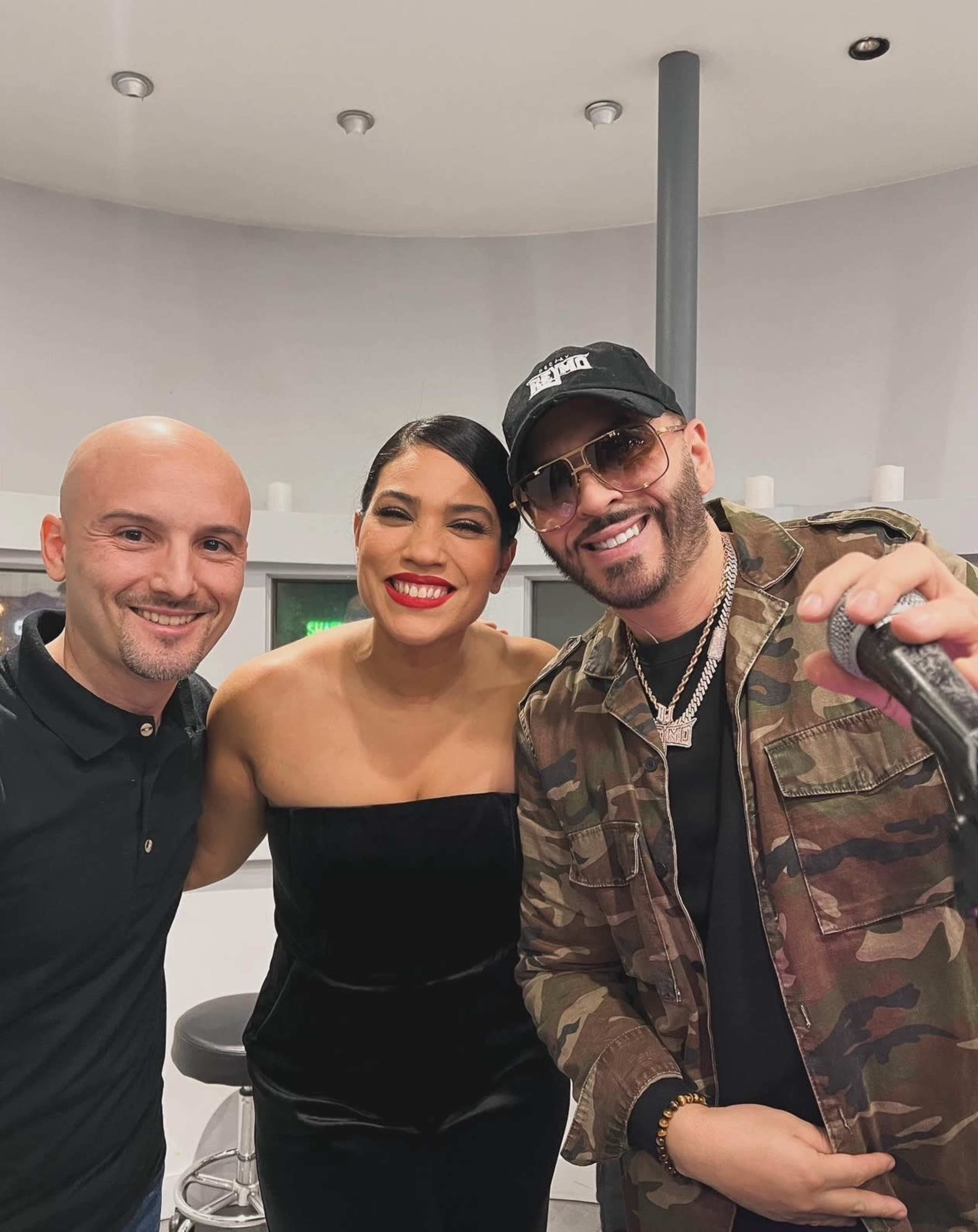 Three individuals pose together for a selfie in an indoor setting, with one wearing a camouflage jacket and sunglasses, the second in a black dress, and the third in a black polo shirt.