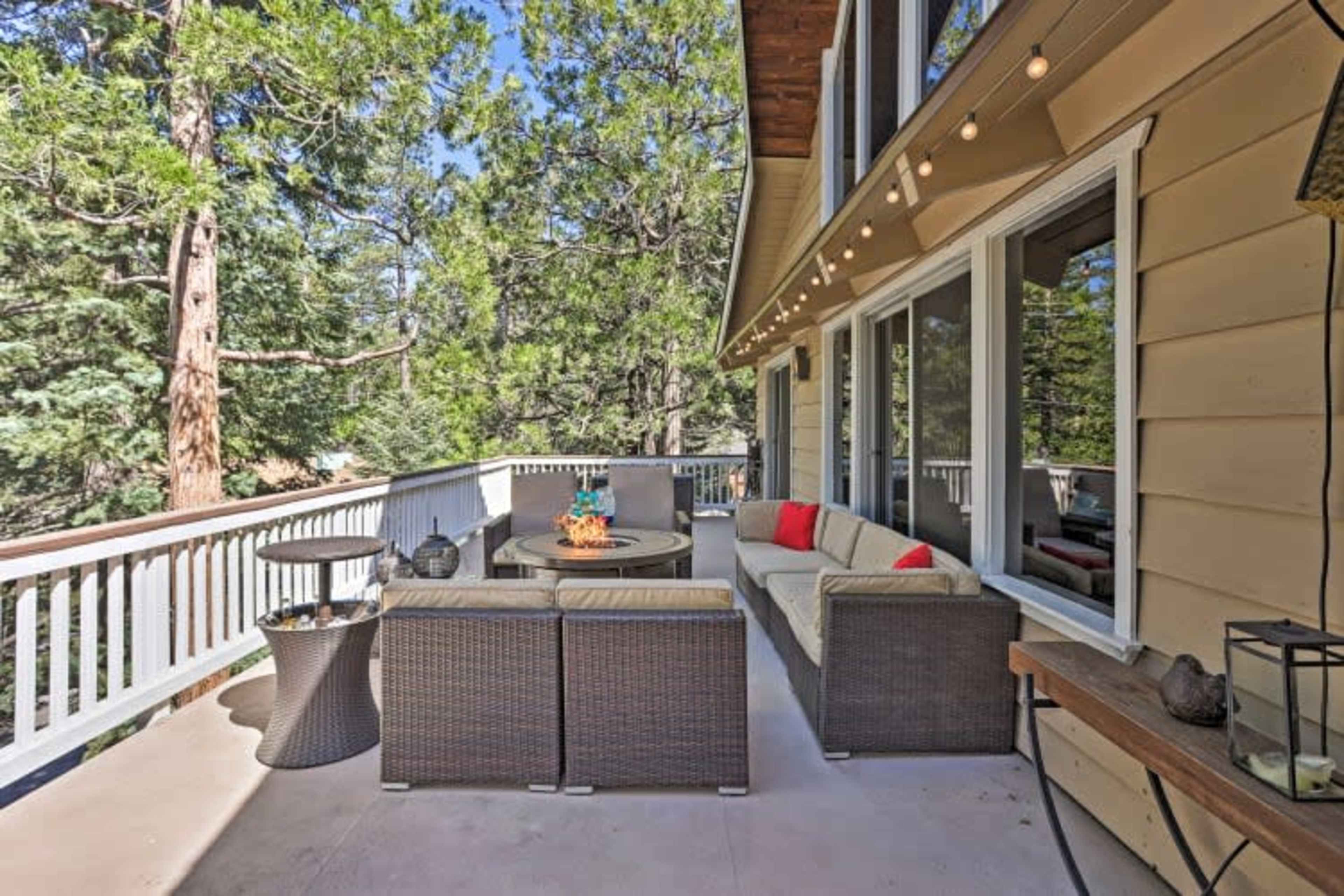 A patio features a seating area with a sectional sofa, a circular table, and decorative pillows, all surrounded by pine trees and string lights.
