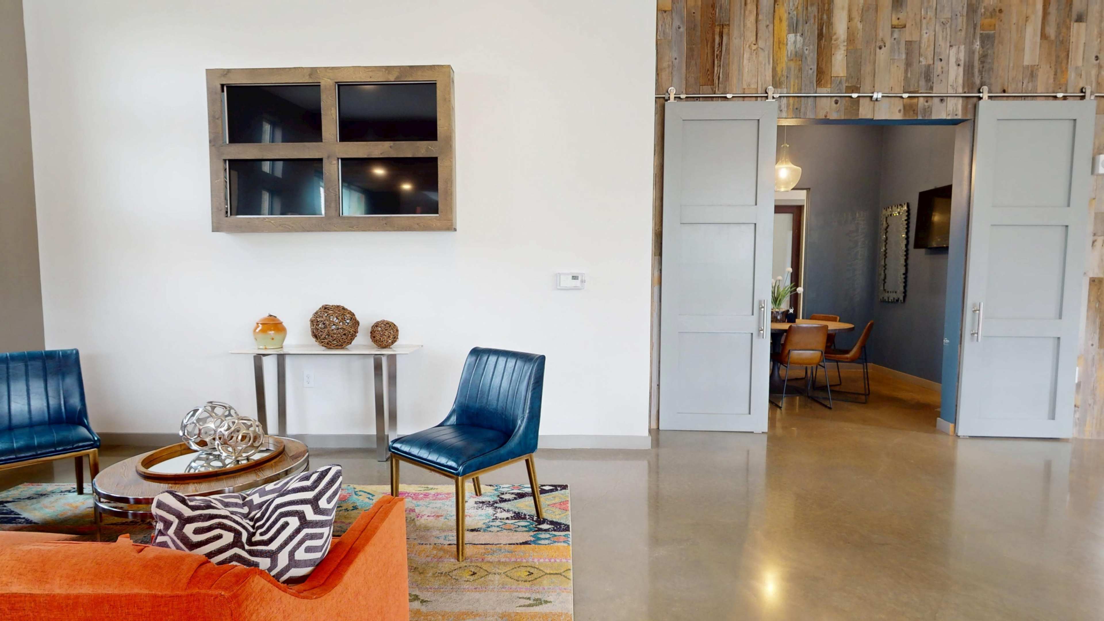 A modern living room features a stylish orange sofa, a round coffee table, and blue chairs, with an open doorway leading to a dining area in the background.