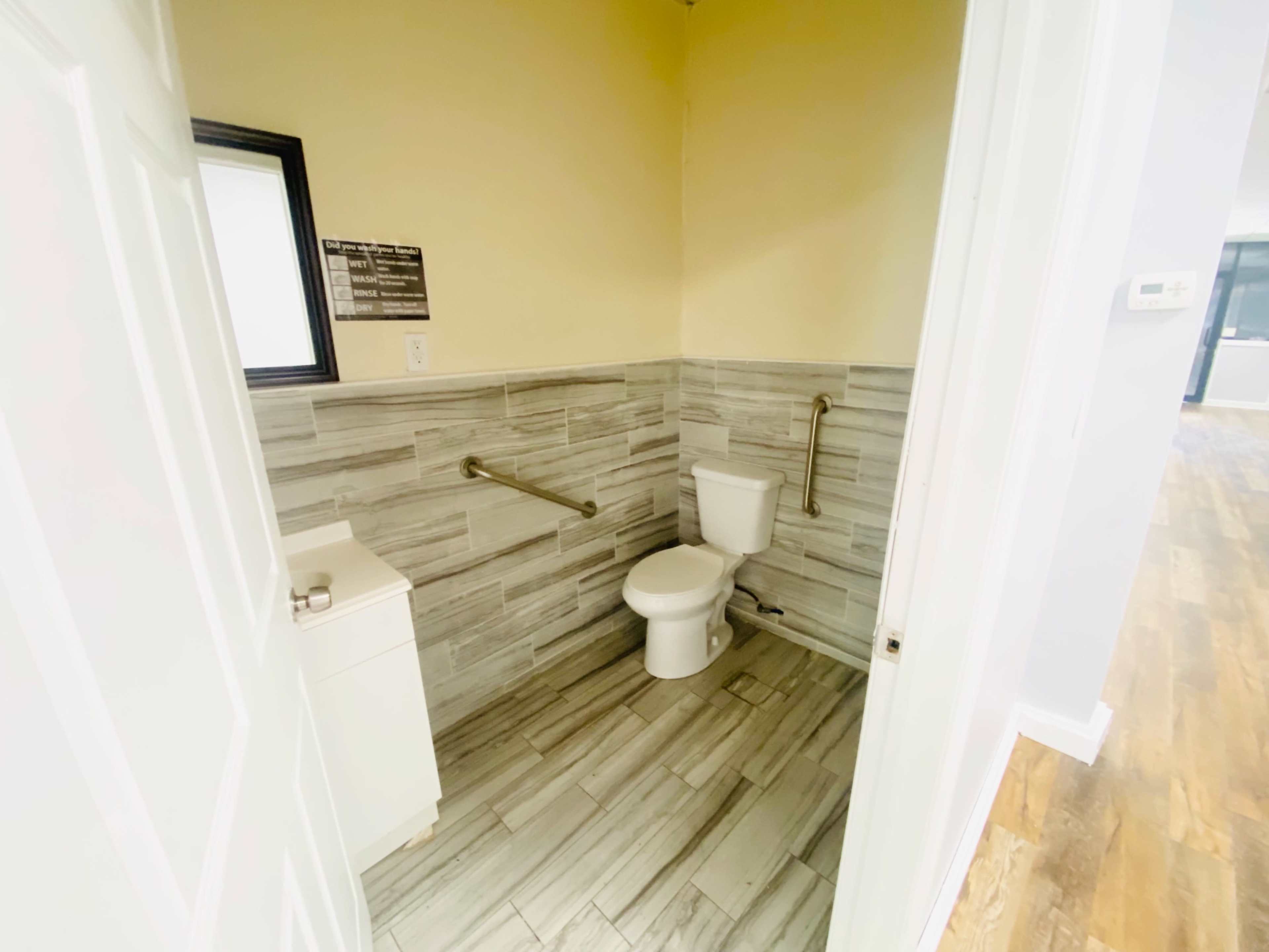 The image shows a small bathroom featuring a toilet, a sink, grab bars, and tiled walls.