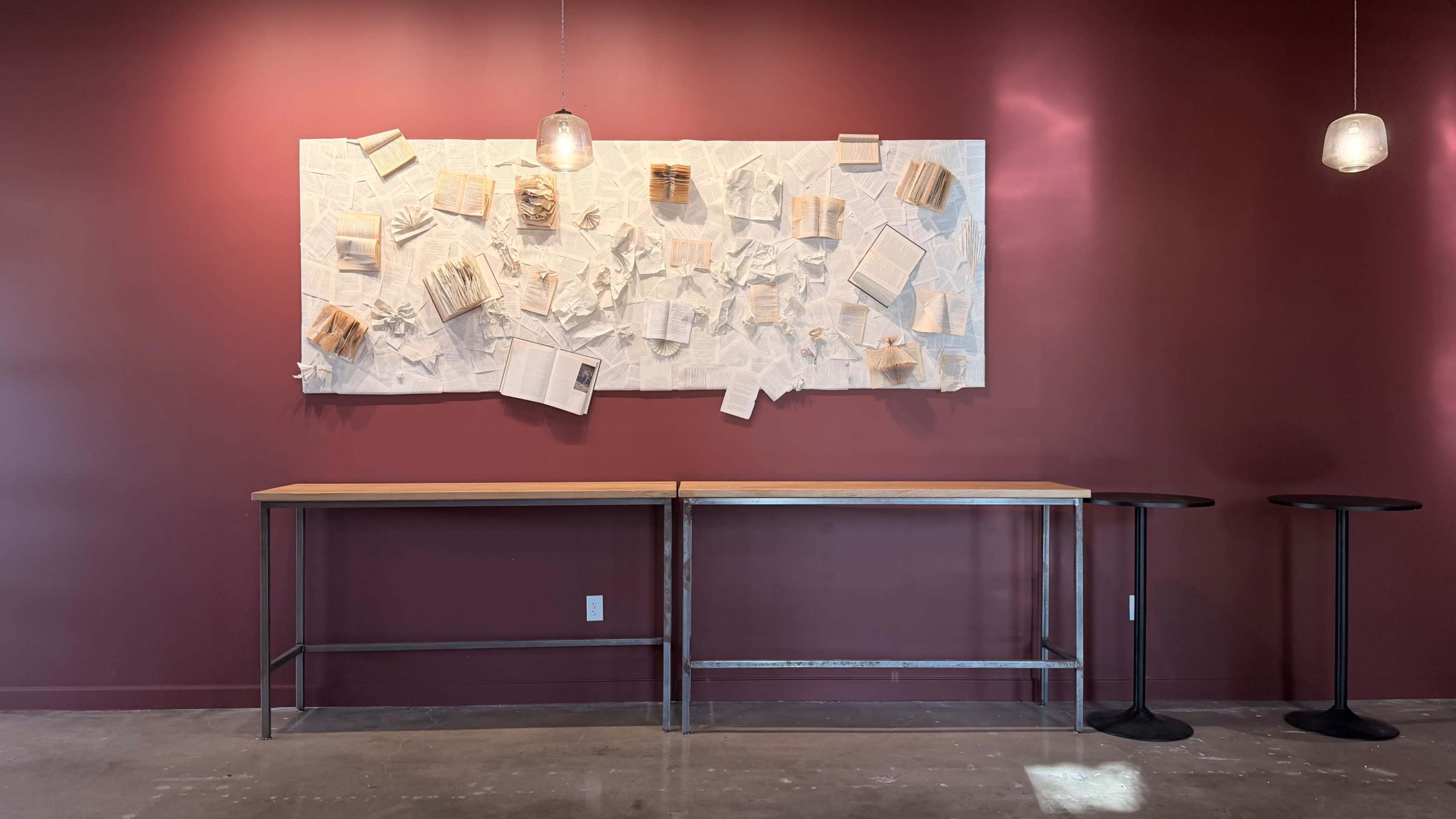 The image shows a minimalist interior with a maroon wall, a wooden table with a metal base, and a wall-mounted art piece featuring various paper elements arranged on textured fabric.