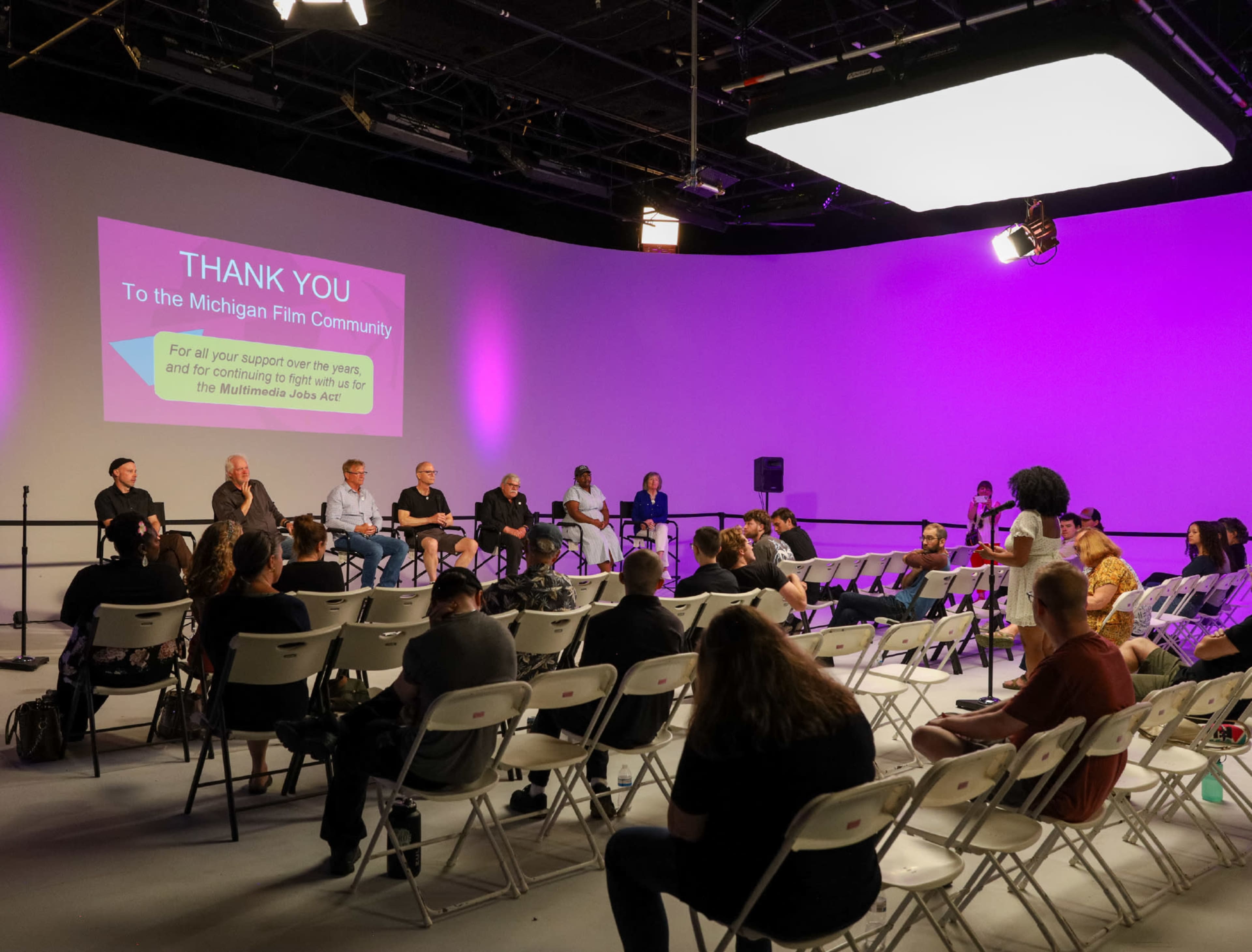 A group of people sits in a studio setting with a large thank-you message displayed on a screen, while a panel discussion takes place at the front.