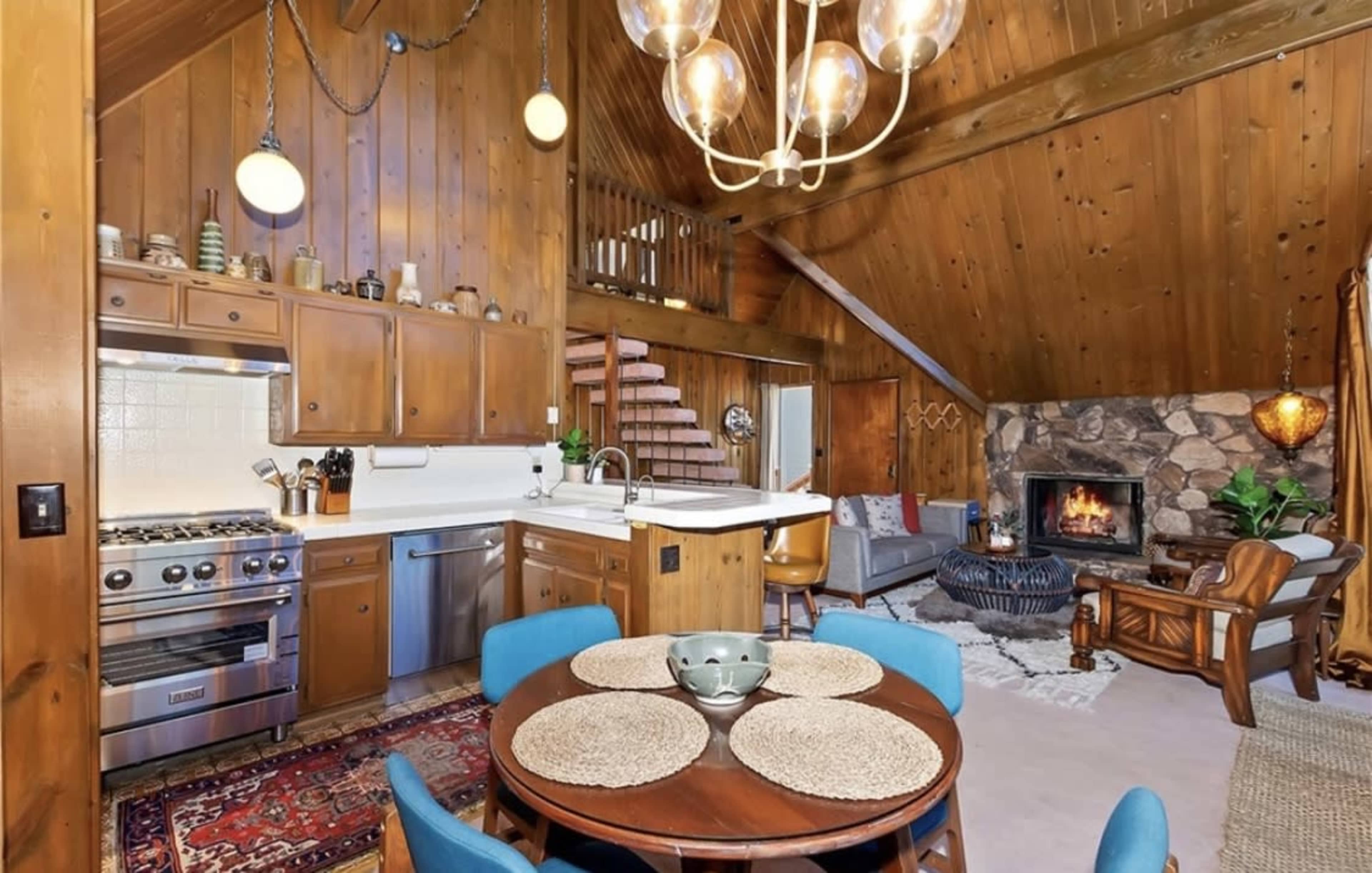 A cozy kitchen and living area with wooden walls, a stone fireplace, a dining table, and stairs leading to a loft.