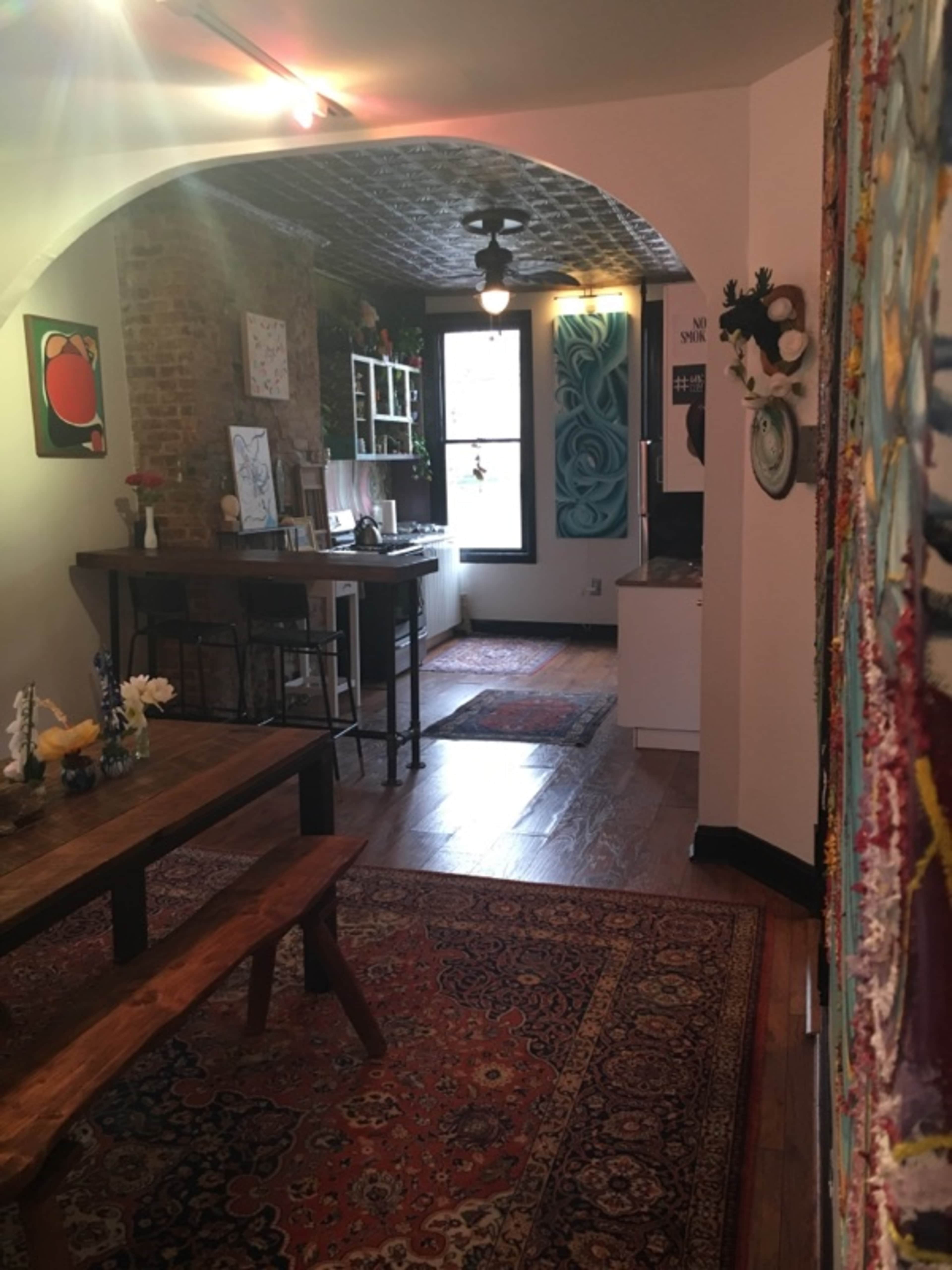 The image shows a cozy kitchen and dining area with wooden furniture, brick walls, and patterned rugs.