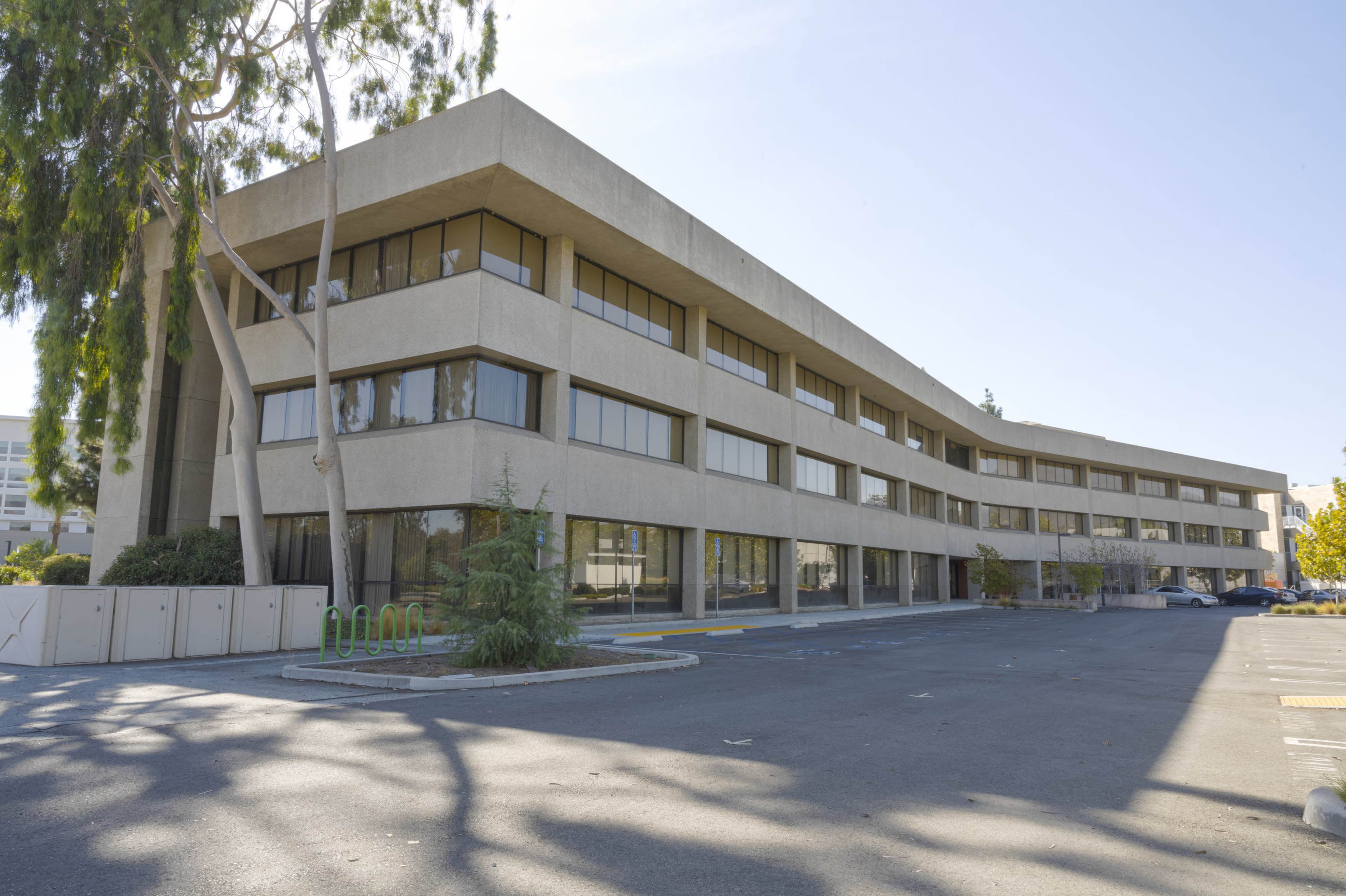 A three-story, modern concrete building with large windows and an adjacent parking area.