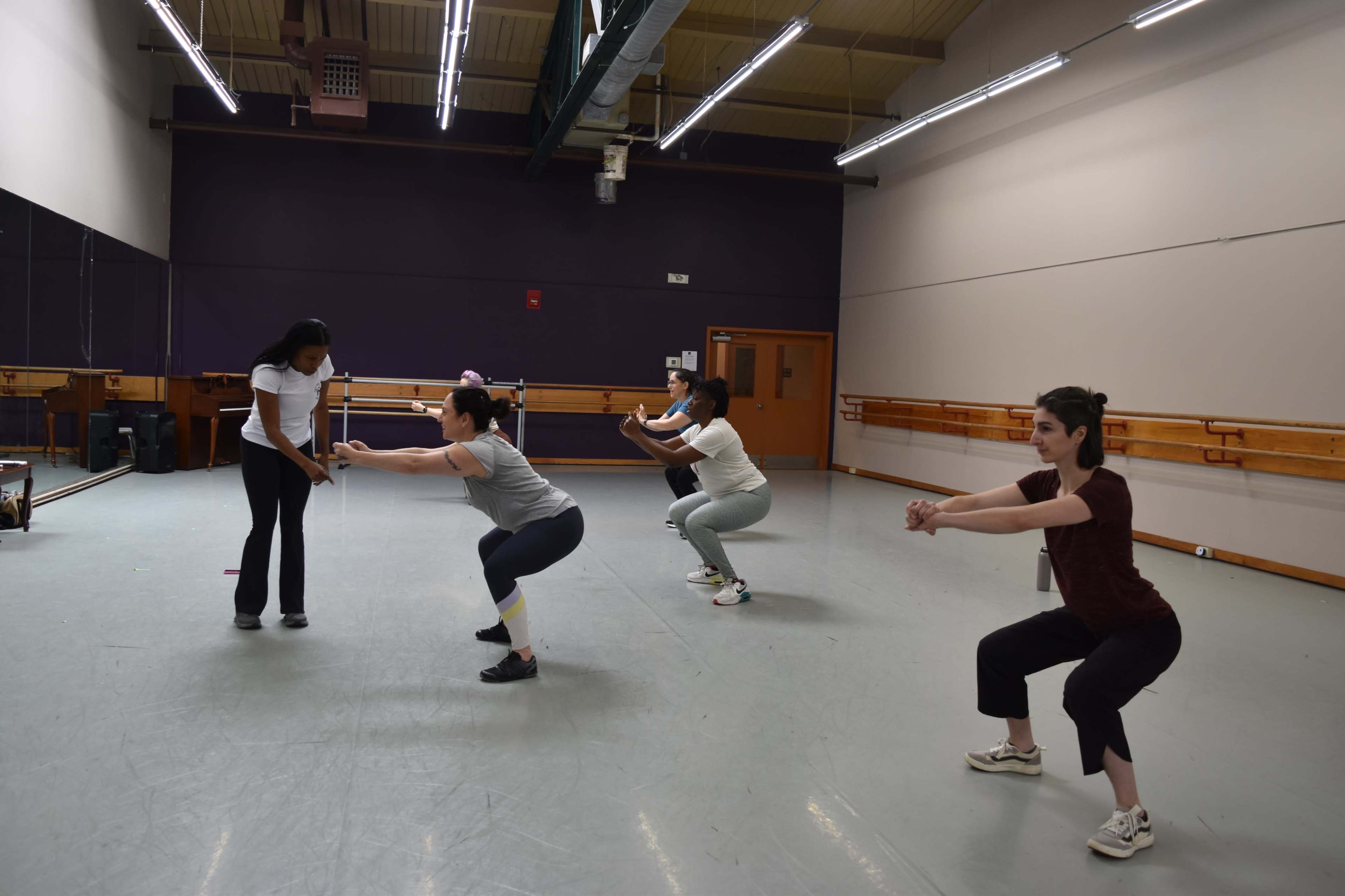 Four individuals are performing squats in a dance studio, with ballet bars visible in the background.