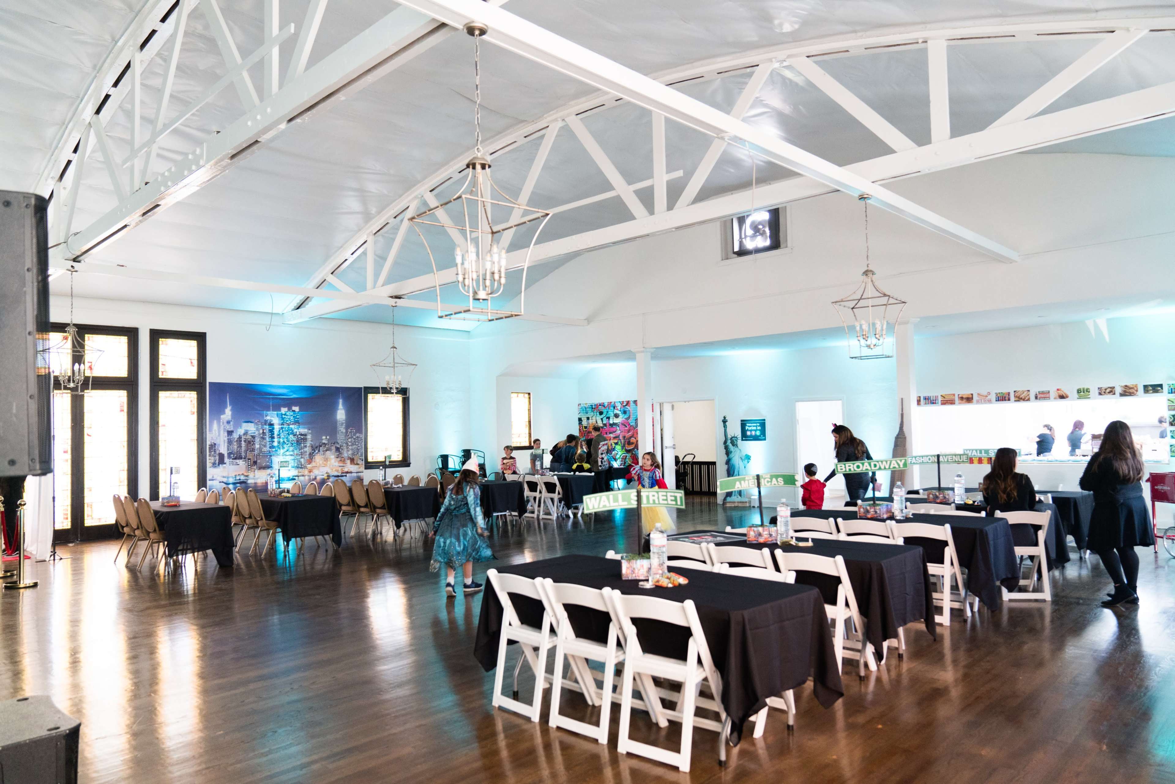 An empty event space with rows of tables set with black tablecloths and white chairs, illuminated by overhead lighting.