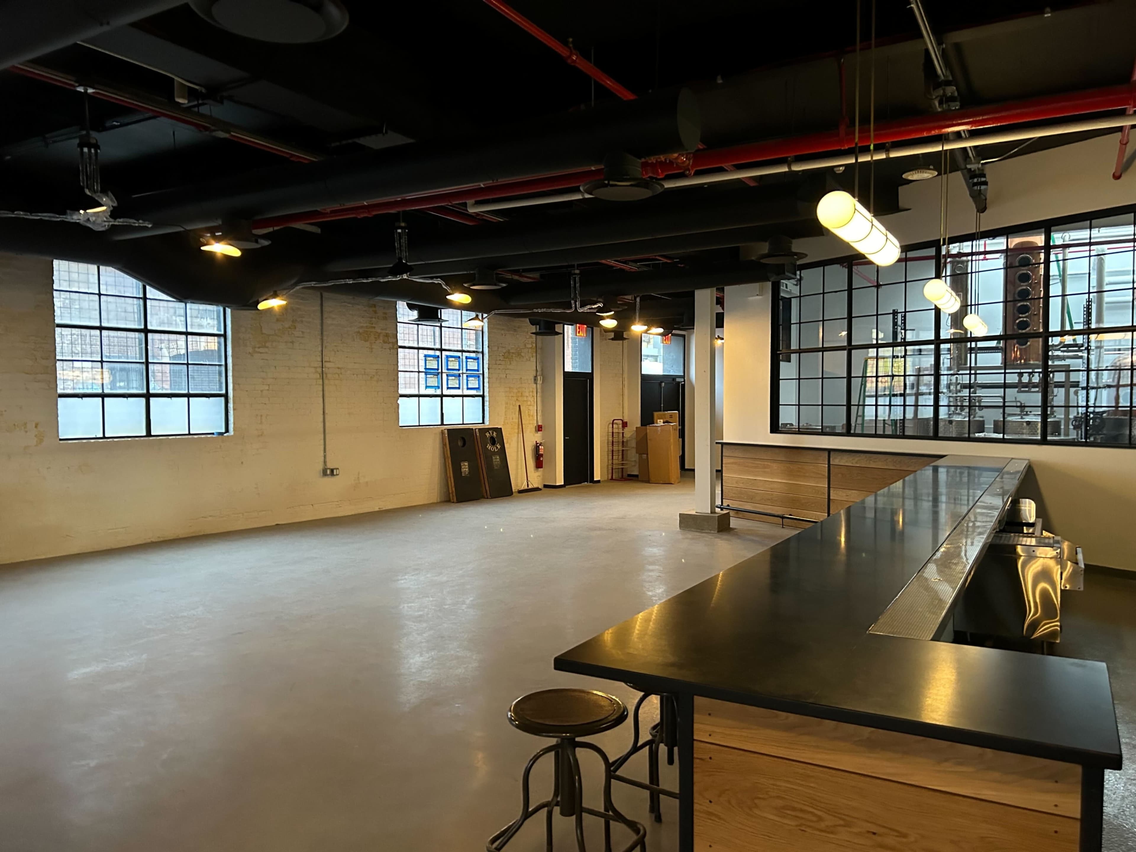 A spacious, empty interior of a room featuring a long bar counter, industrial-style windows, and exposed pipes on the ceiling.